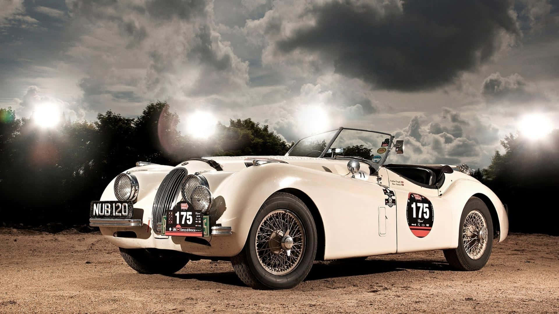 1950年捷豹XK 120古董車圖 壁紙 — 一架編號175的純白色經典賽車，在明亮的聚光燈照射下，戲劇性的多雲天空下，停泊在泥地賽道上。