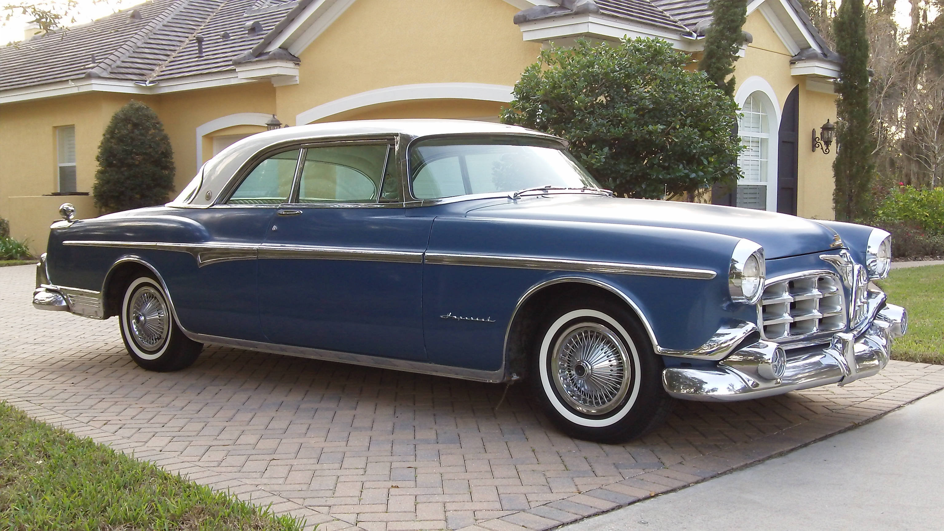 1955 Chrysler Imperial 兩門車 壁紙 — 一架經典藍白色1950年代轎車在日光下閃耀，停泊在黃色房屋前的磚砌車道上。