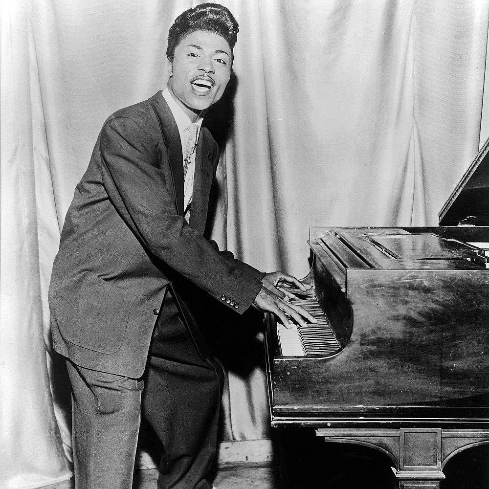 1956年小理查德黑白舞台彈鋼琴演唱 壁紙 — 一位身穿西裝的快樂黑人男子在表演中俯身於三角鋼琴上，背景是垂簾。