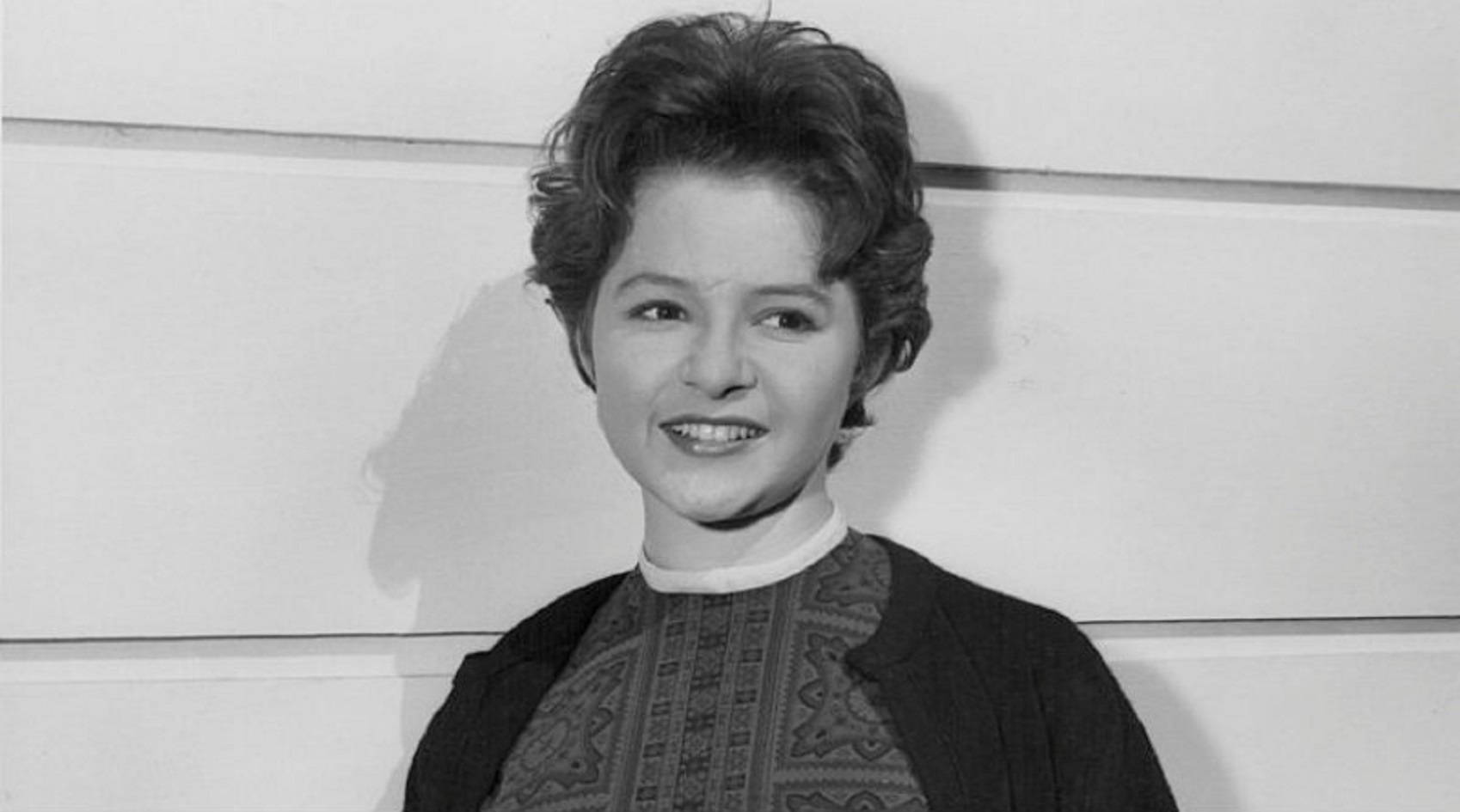 1960年代美國獨唱歌手Brenda Lee黑白寫真 壁紙 — 一幅黑白肖像，一位短曲髮、面帶微笑的女性，身穿圖案上衣和開襟毛衣，靠著嵌板牆。