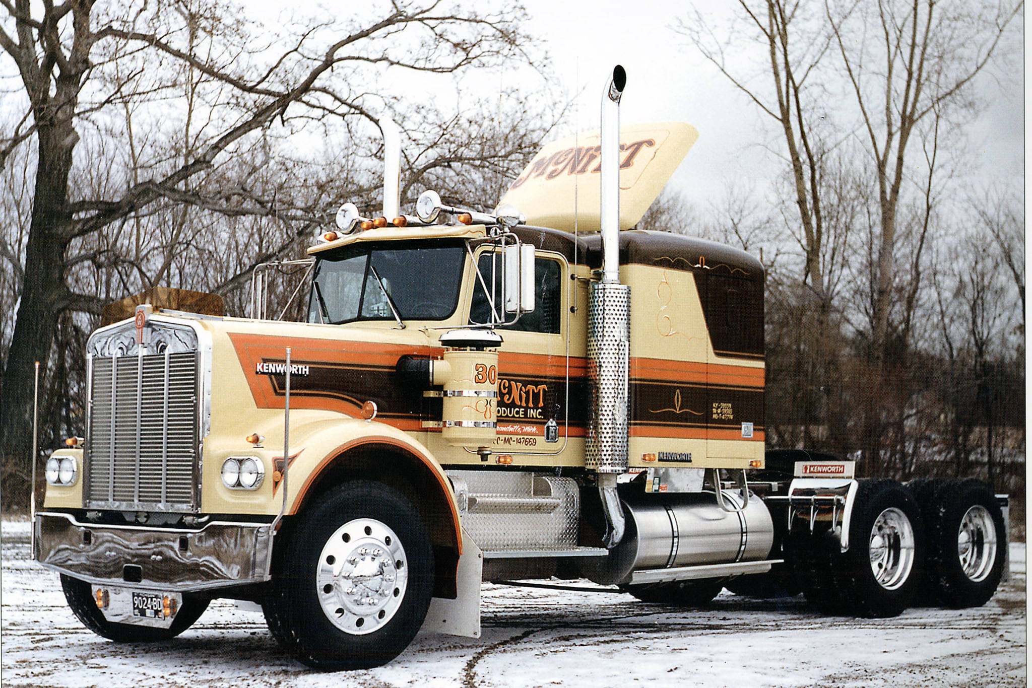 1961 年肯沃斯 W900 壁紙 — 一輛奶油色和棕色的經典Kenworth半掛卡車，帶有鍍鉻裝飾，停在白雪覆蓋的森林空地上。