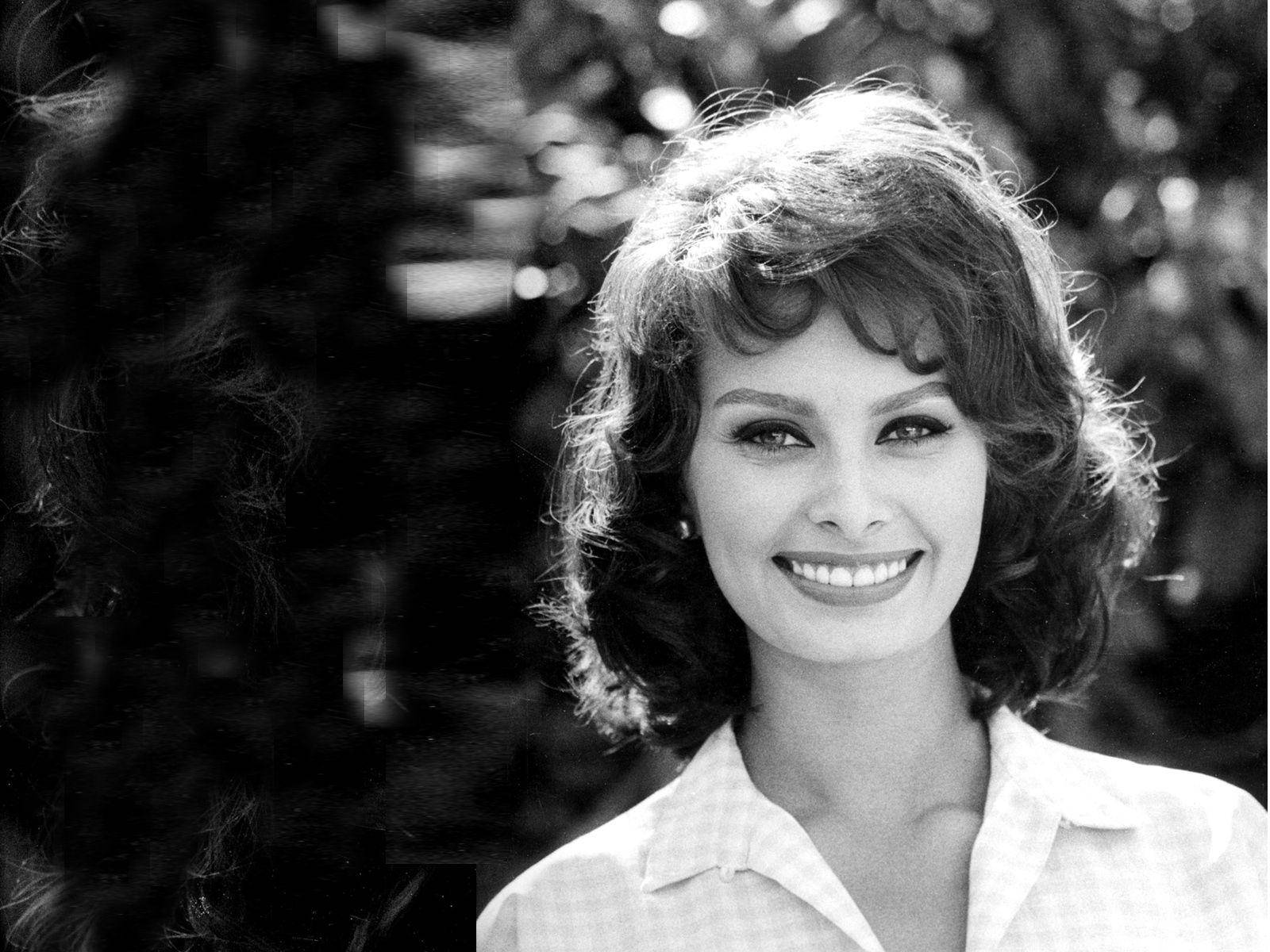 1964年光彩照人的索菲亞·羅蘭 壁紙 — 一位光彩照人的女性，擁有豐盈的捲髮，在經典黑白背景下，對著柔和模糊的樹葉背景溫暖微笑。