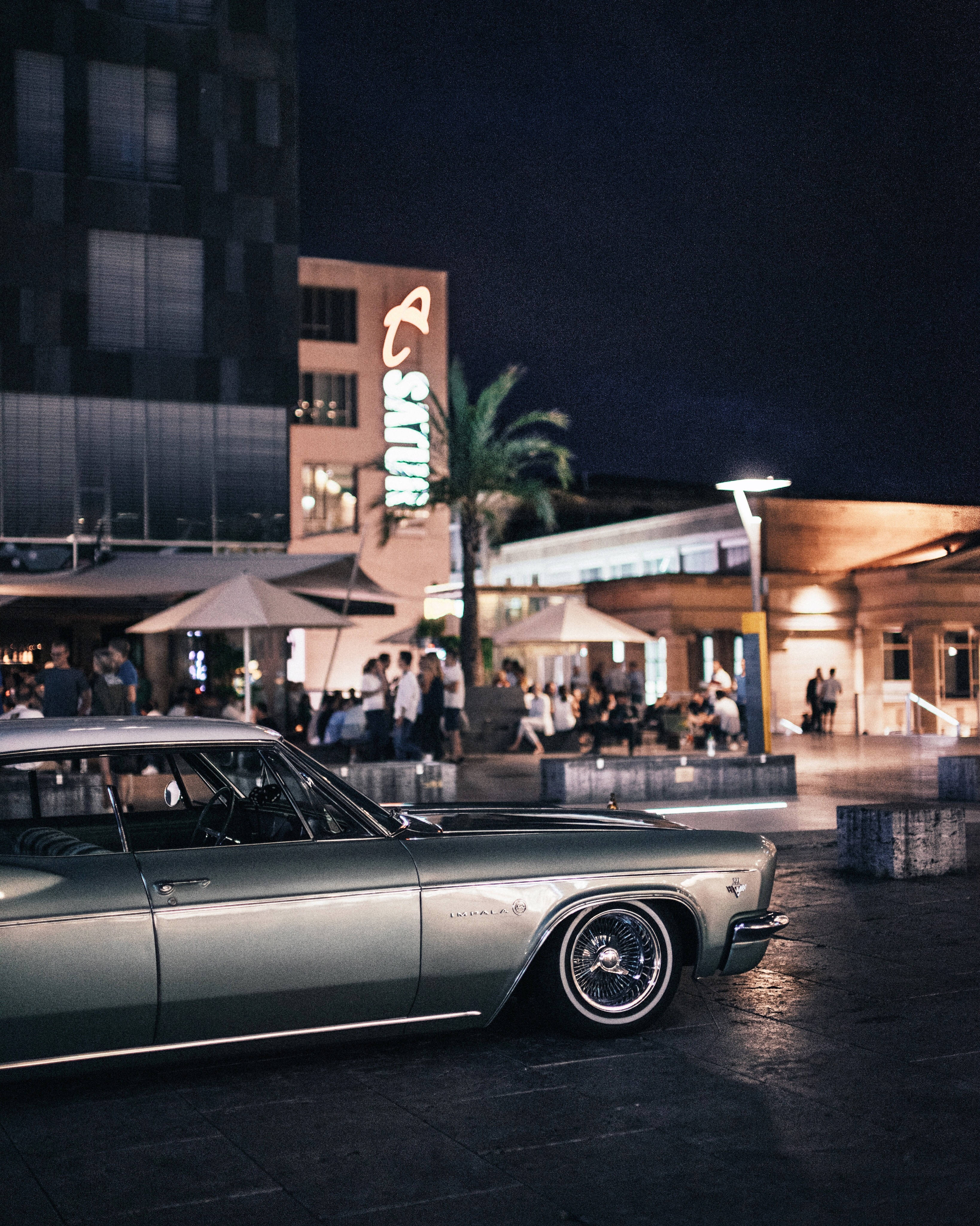 1965銀色低 Rider雪佛蘭Impala 壁紙 — 一輛復古銀色Impala在霓虹燈下閃耀，夜晚的繁華城市廣場上，人們在遮陽傘下用餐。