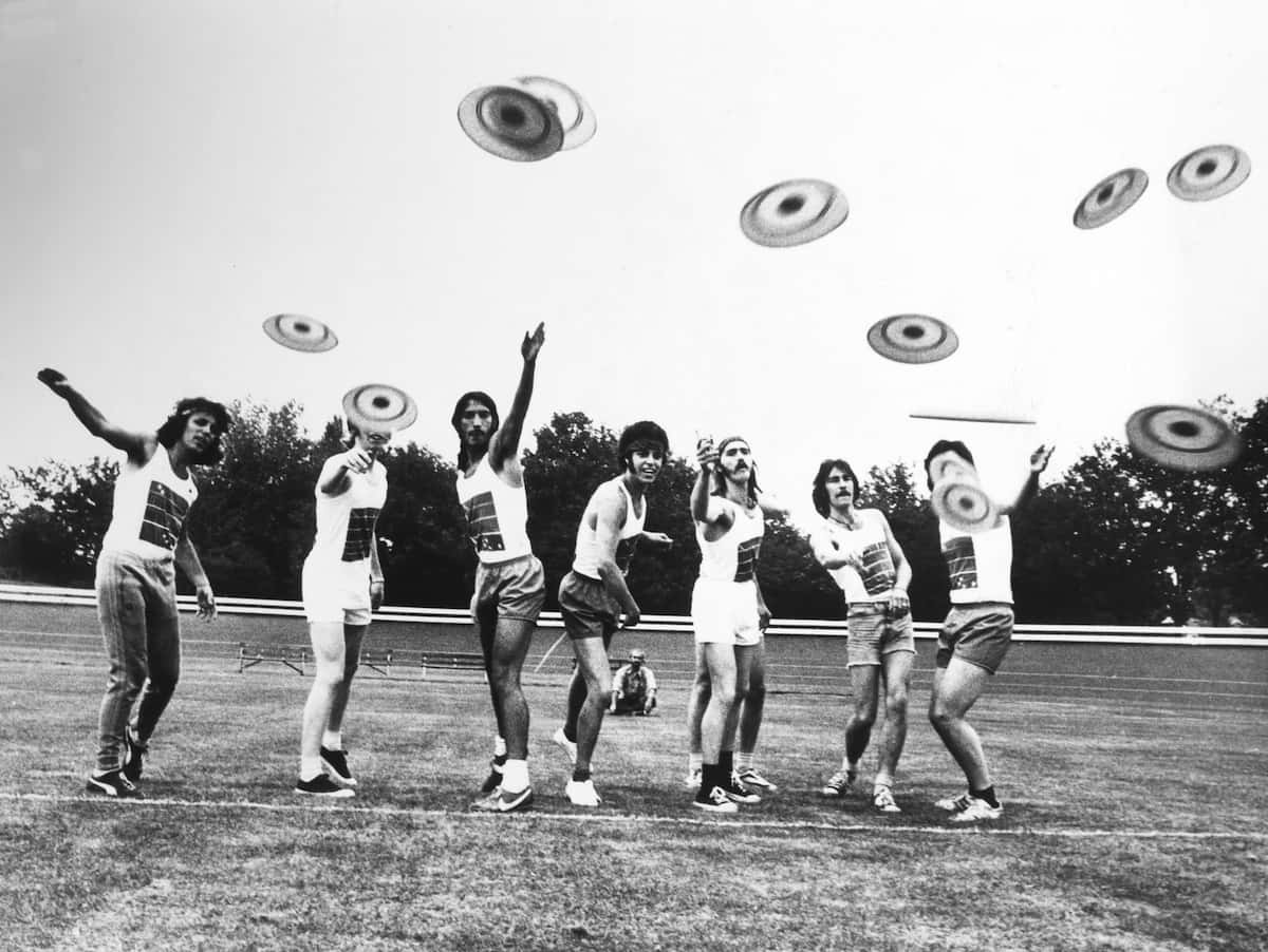 1966 年英國最佳 Ultimate Frisbee 背景 壁紙 — 七名身穿復古運動服的男士在草地上同時擲飛盤，背景有樹木。
