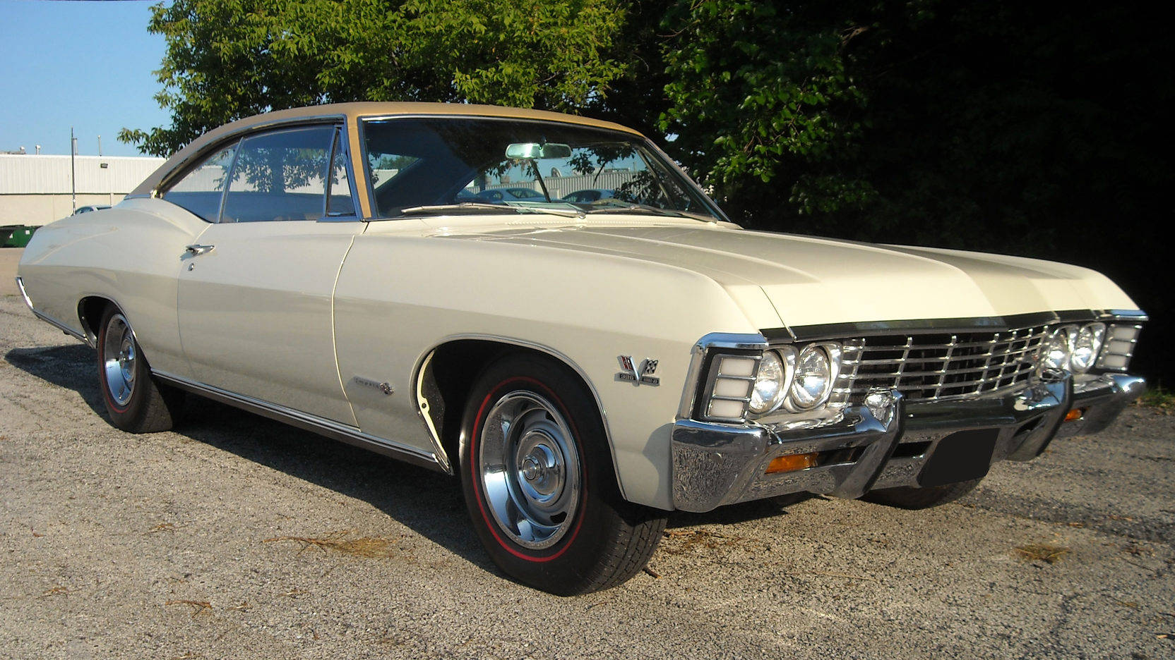 復古美學 - 1967年奶油色雪佛蘭Impala 壁紙 — 一輛奶油色經典肌肉車，帶有鍍鉻裝飾和紅線輪胎，停在明亮的日光下的礫石上。