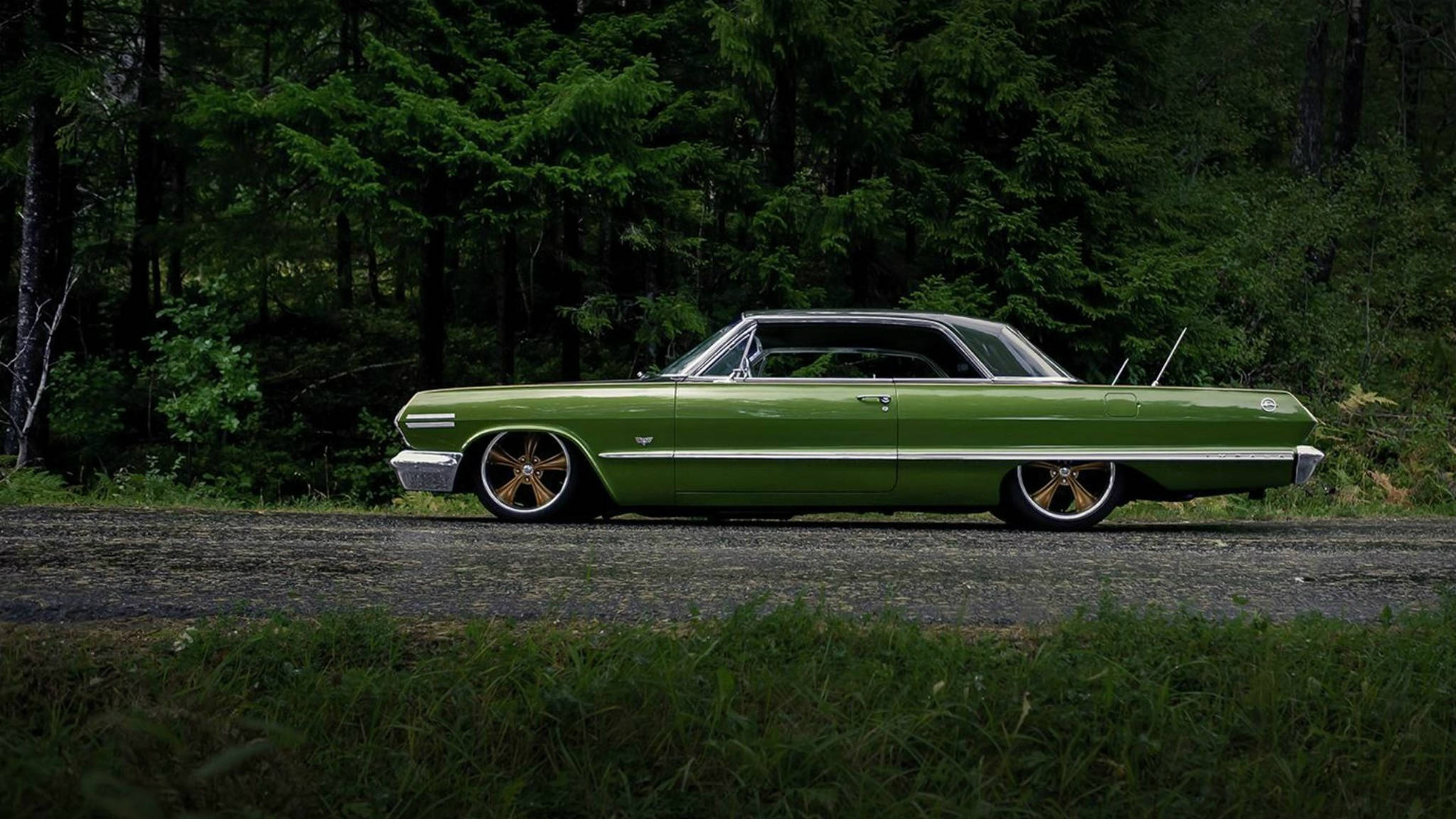 1967年綠色雪佛蘭 Impala 壁紙 — 一架復古綠色肌肉車配金色輪圈，停泊在被茂密森林包圍的碎石路上。