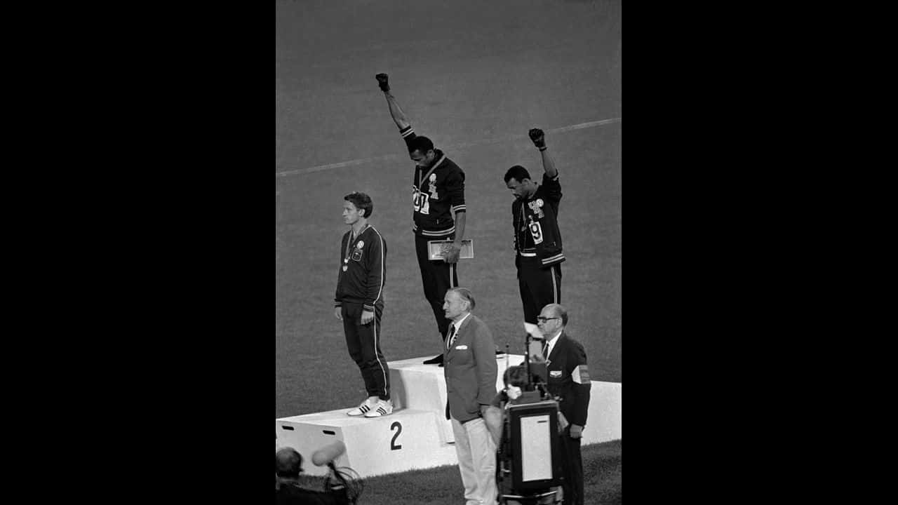 1968 年奧運會黑人權力舉手致敬 壁紙 — 三位運動員站在頒獎台上，一人舉起拳頭抗議，另一人在獎牌儀式中莊嚴站立。