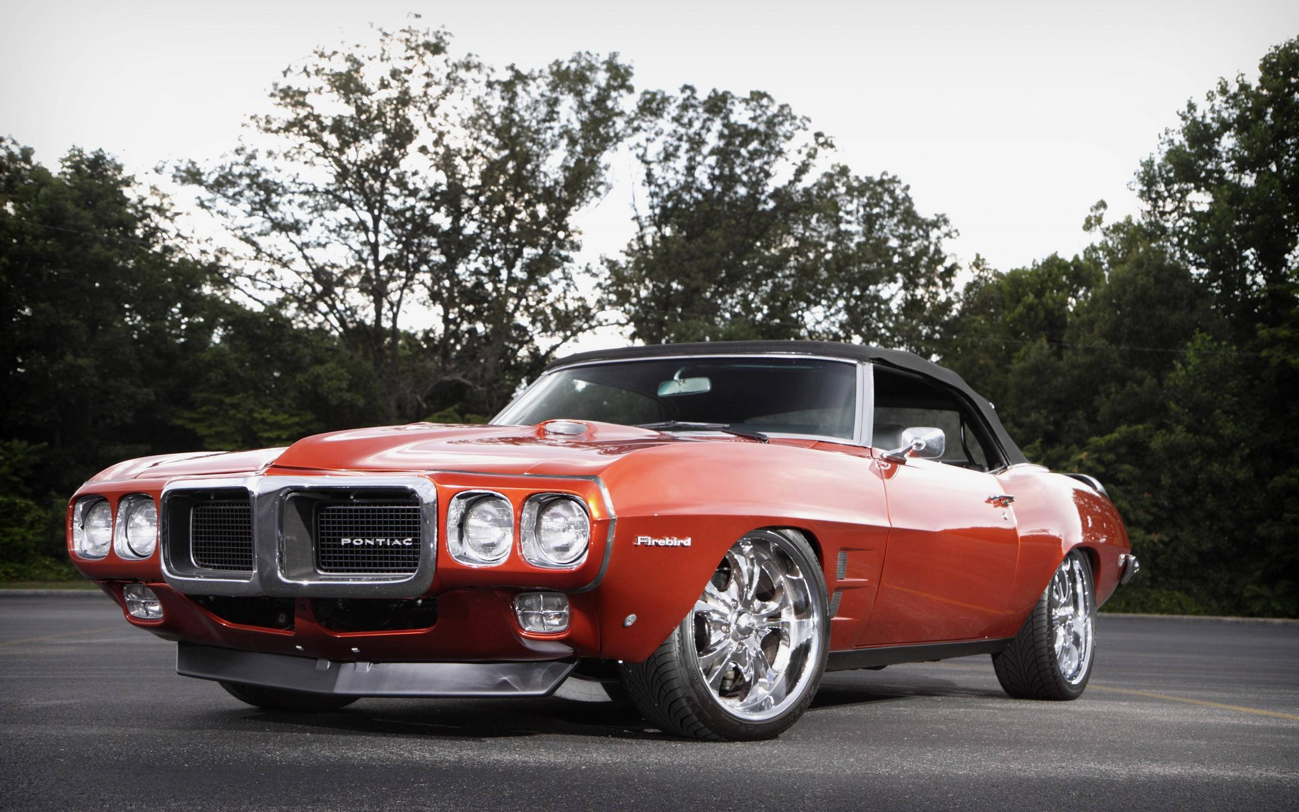 1969年橙色龐蒂克火鳥 壁紙 — 一輛充滿活力的橙色Pontiac Firebird開篷車，配備鍍鉻輪圈，停在柏油路上，周圍綠樹成蔭。