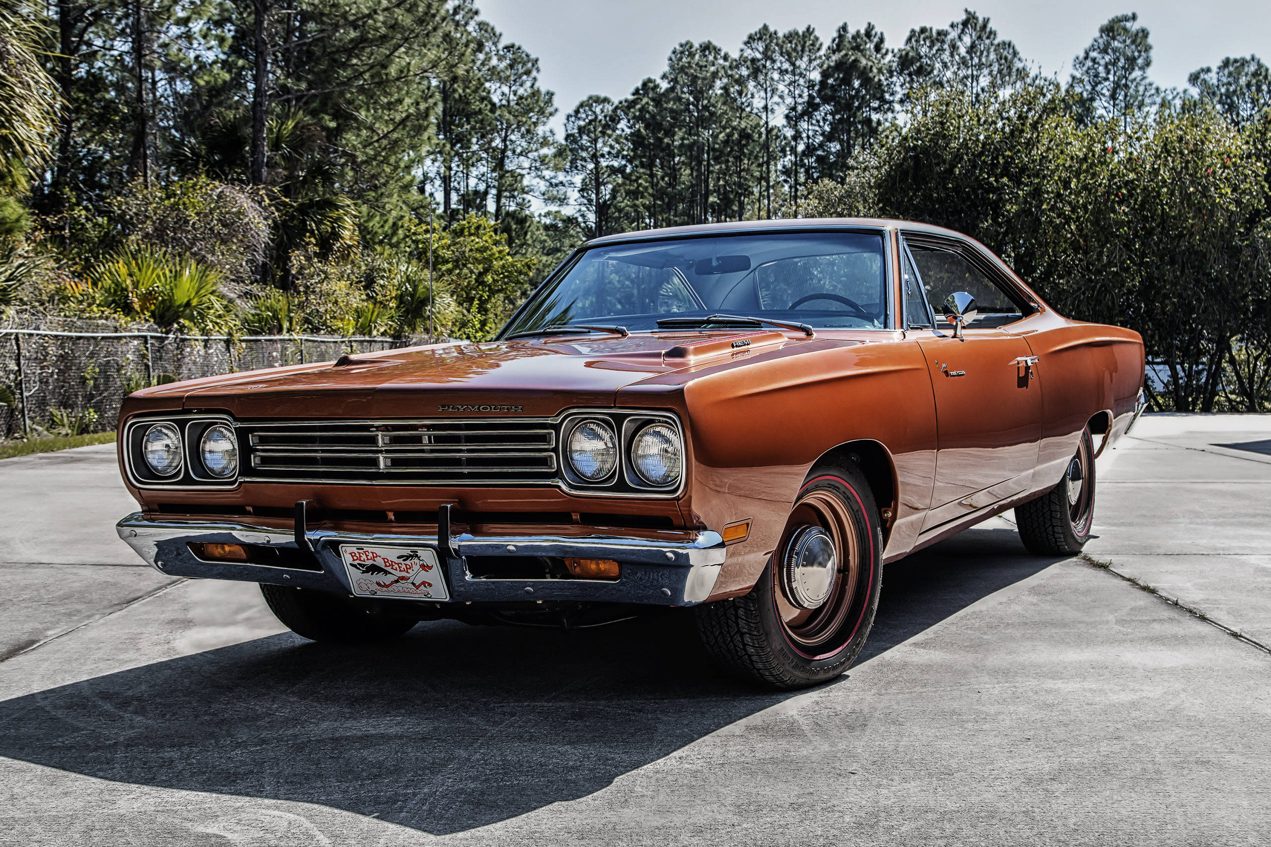 經典 1969 龐蒂克Road Runner，引人注目的棕色車身 壁紙 — 一輛復古橙色道奇Dart肌肉車在明亮的陽光下閃閃發光，停泊在由高大松樹環繞的混凝土車道上。