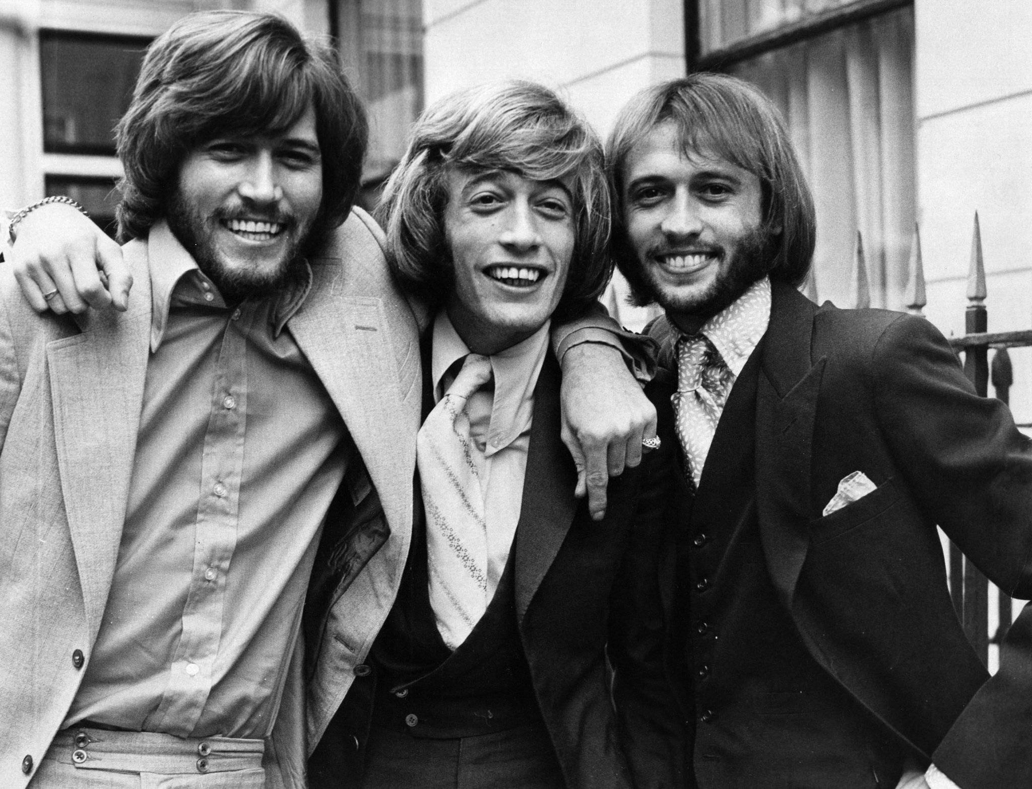 比吉斯1970年倫敦黑白照 壁紙 — 三位留長髮、穿著1970年代時尚的 smiling 男士在戶外黑白照片中緊密擺姿勢。