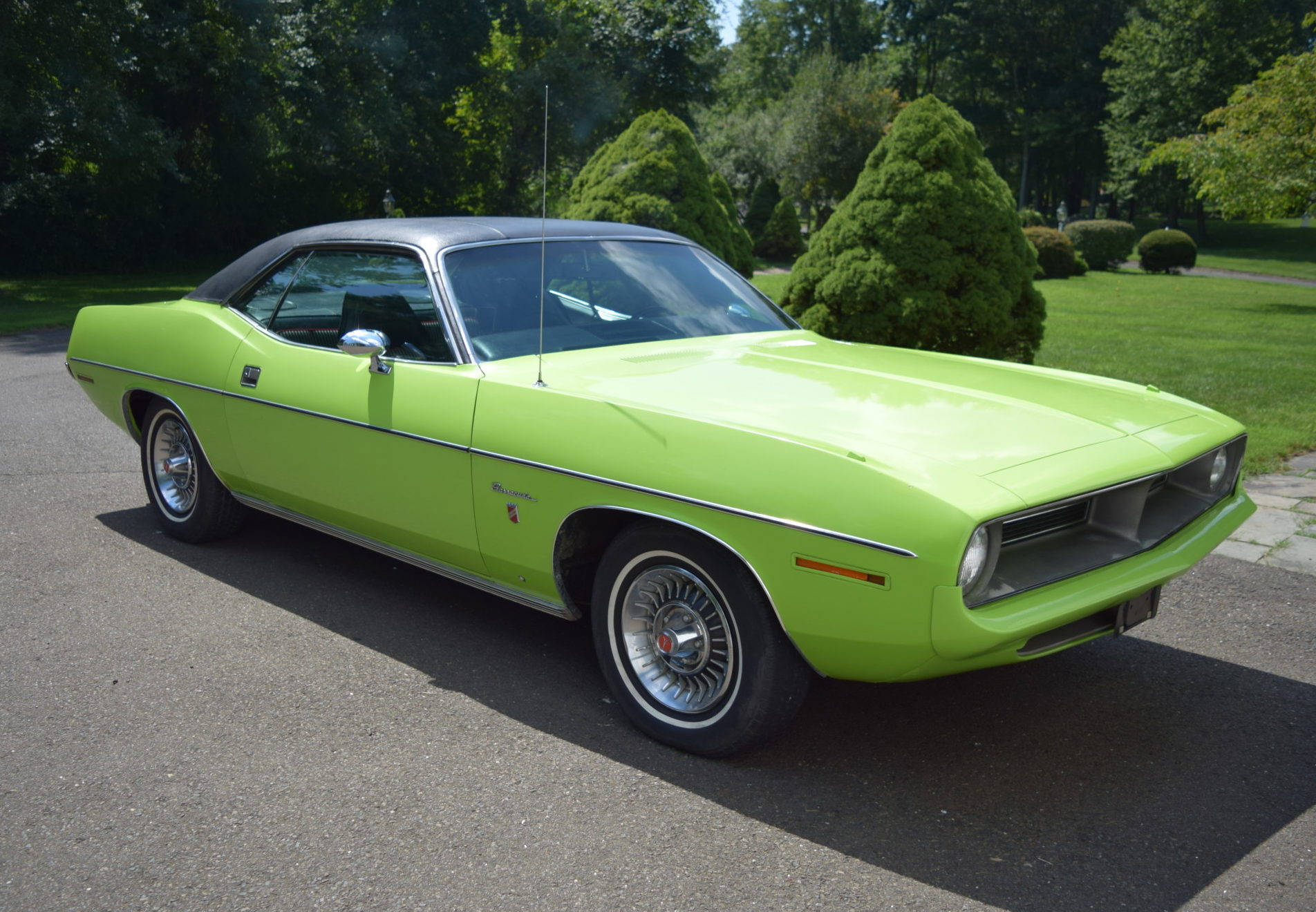 1970 年綠色普利茅斯 Barracuda 壁紙 — 一輛充滿活力的青檸綠色經典肌肉車，黑色車頂，停在瀝青路面上，在明亮的日光下，周圍環繞著茂密的綠樹和修剪整齊的灌木叢。