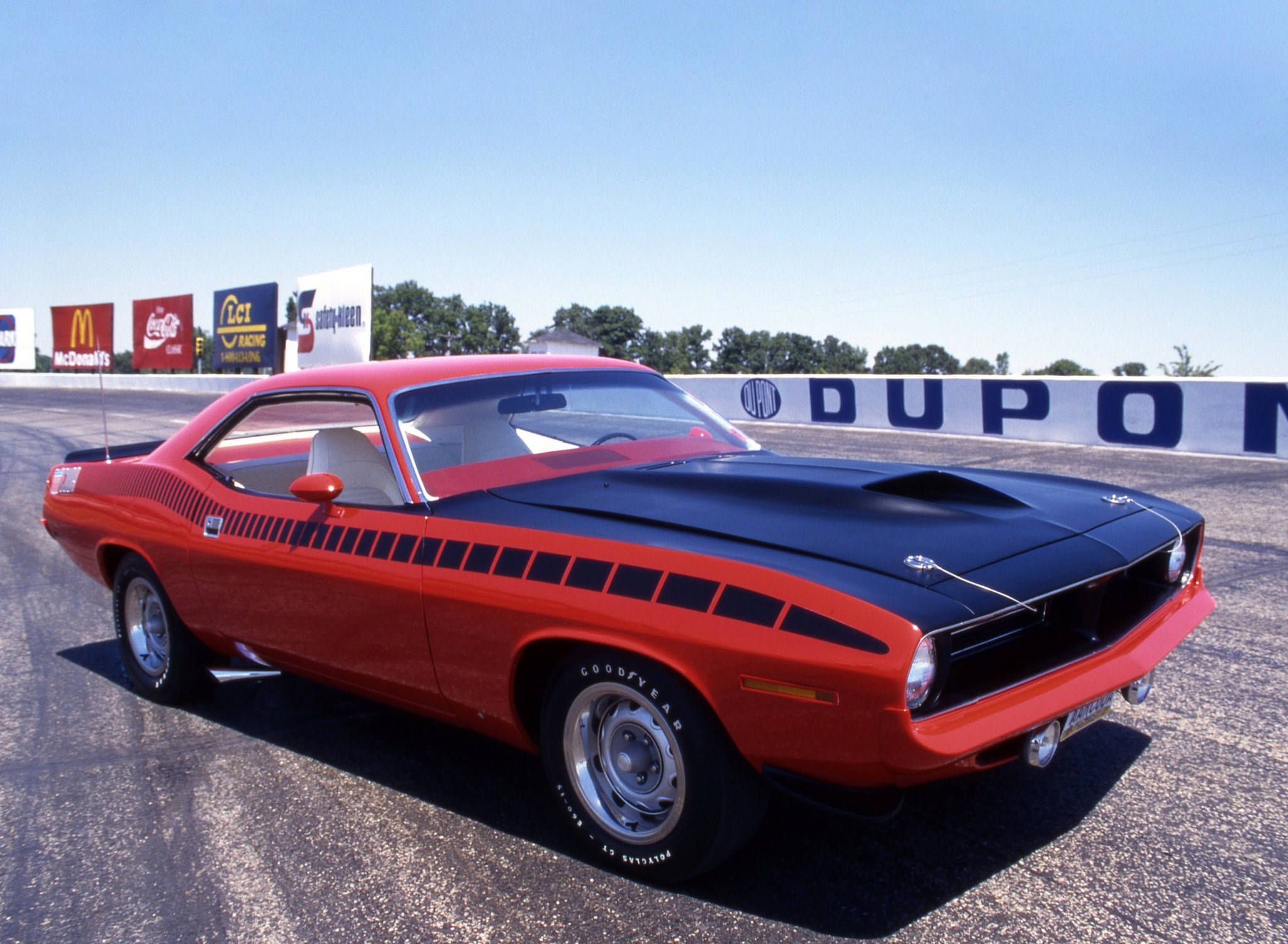 1970年款紅黑配色Plymouth Barracuda 壁紙 — 一輛大膽的紅黑經典肌肉車，帶有黑色賽車條紋，停在陽光明媚的賽車場上，背景有贊助商標誌。