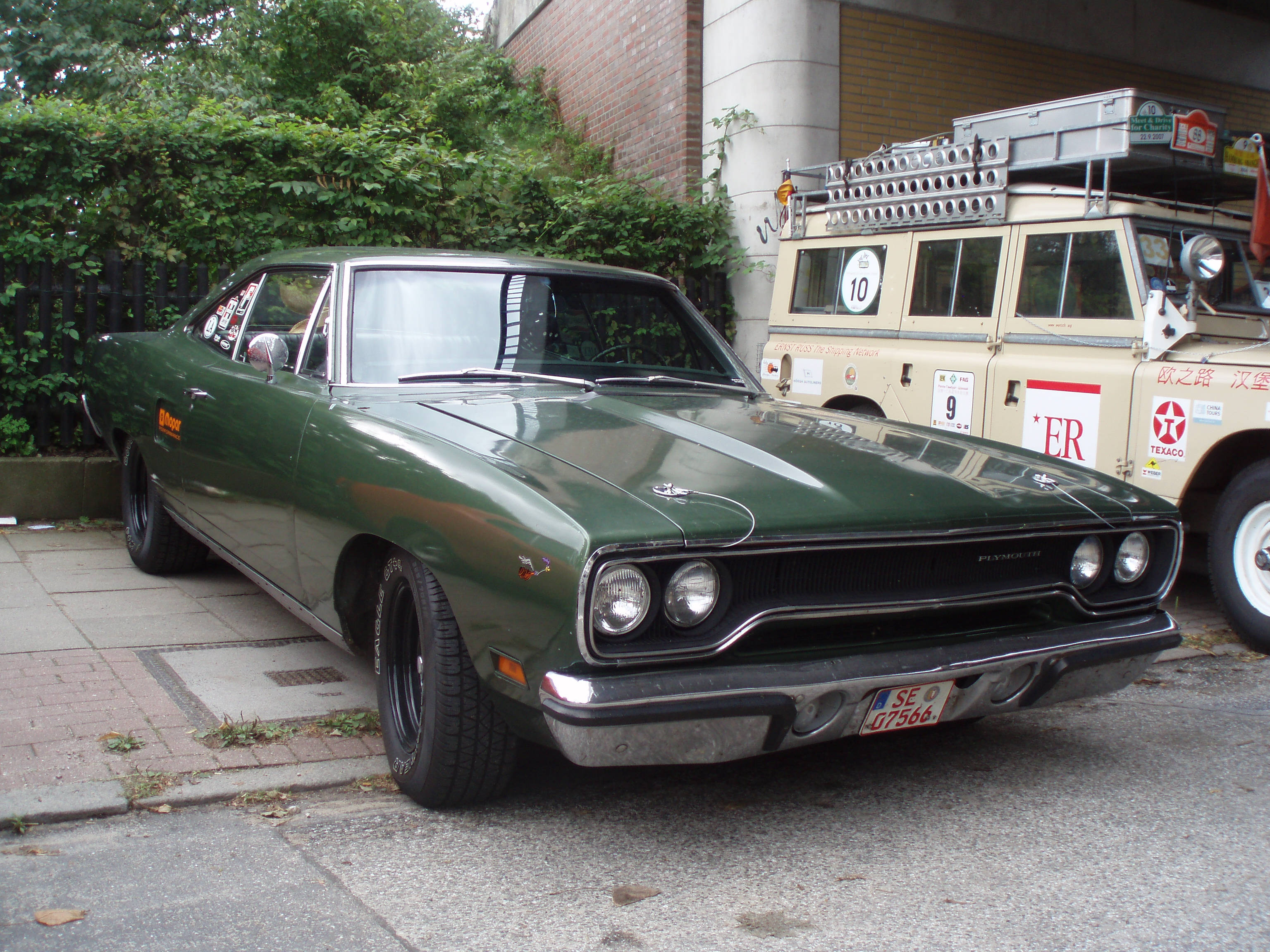 1970年款Plymouth Road Runner深綠色 壁紙 — 一架復古綠色Dodge Dart肌肉車停泊在人行道上，旁邊是一輛帶有賽車貼紙的米色賽車卡車。