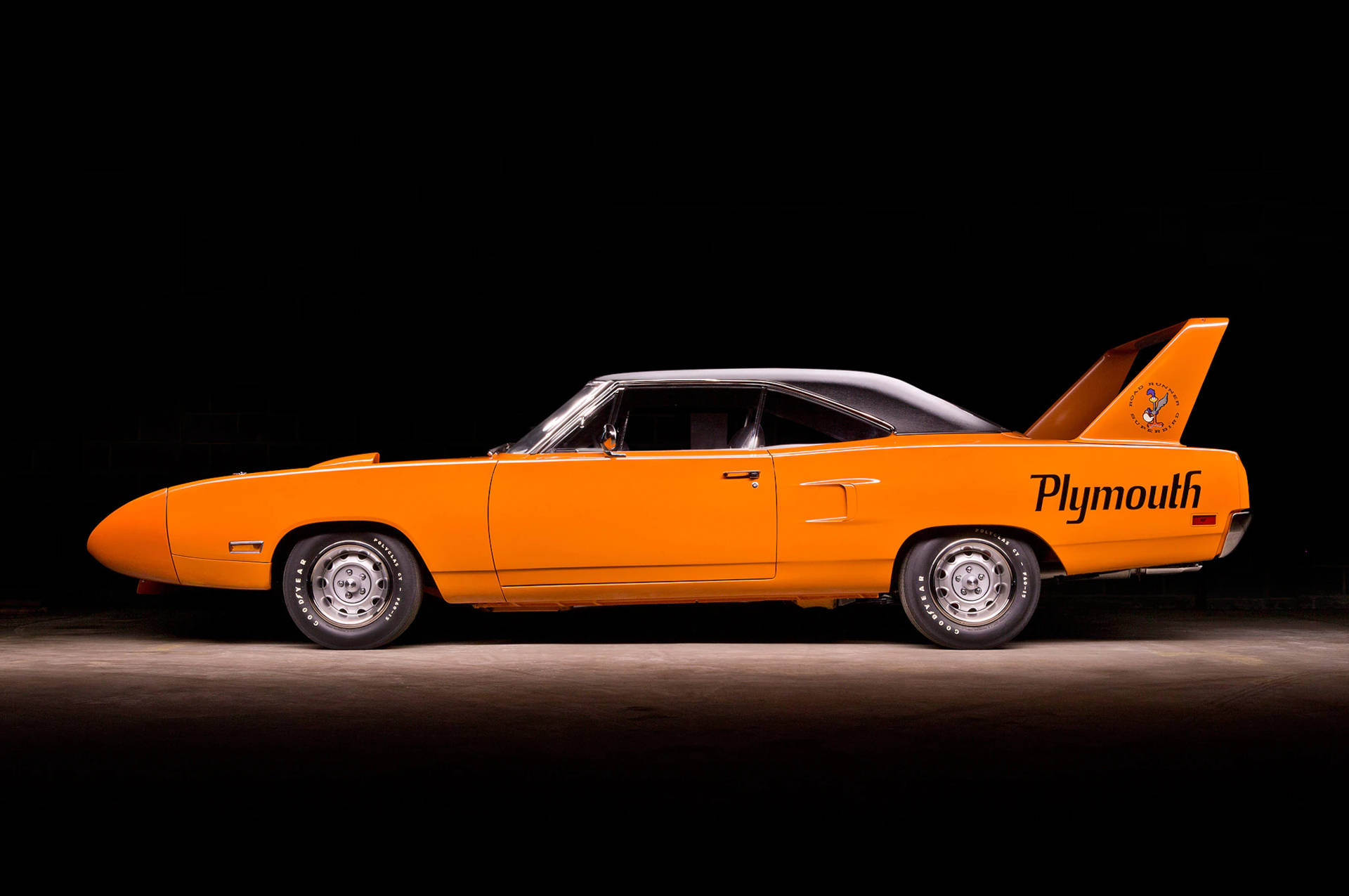 1970年款Plymouth Superbird搶眼橙色 壁紙 — 一架充滿活力的橙色1960年代Plymouth Superbird肌肉車，配備黑色車頂和標誌性尾翼，在黑色背景下的戲劇性工作室燈光中展現。