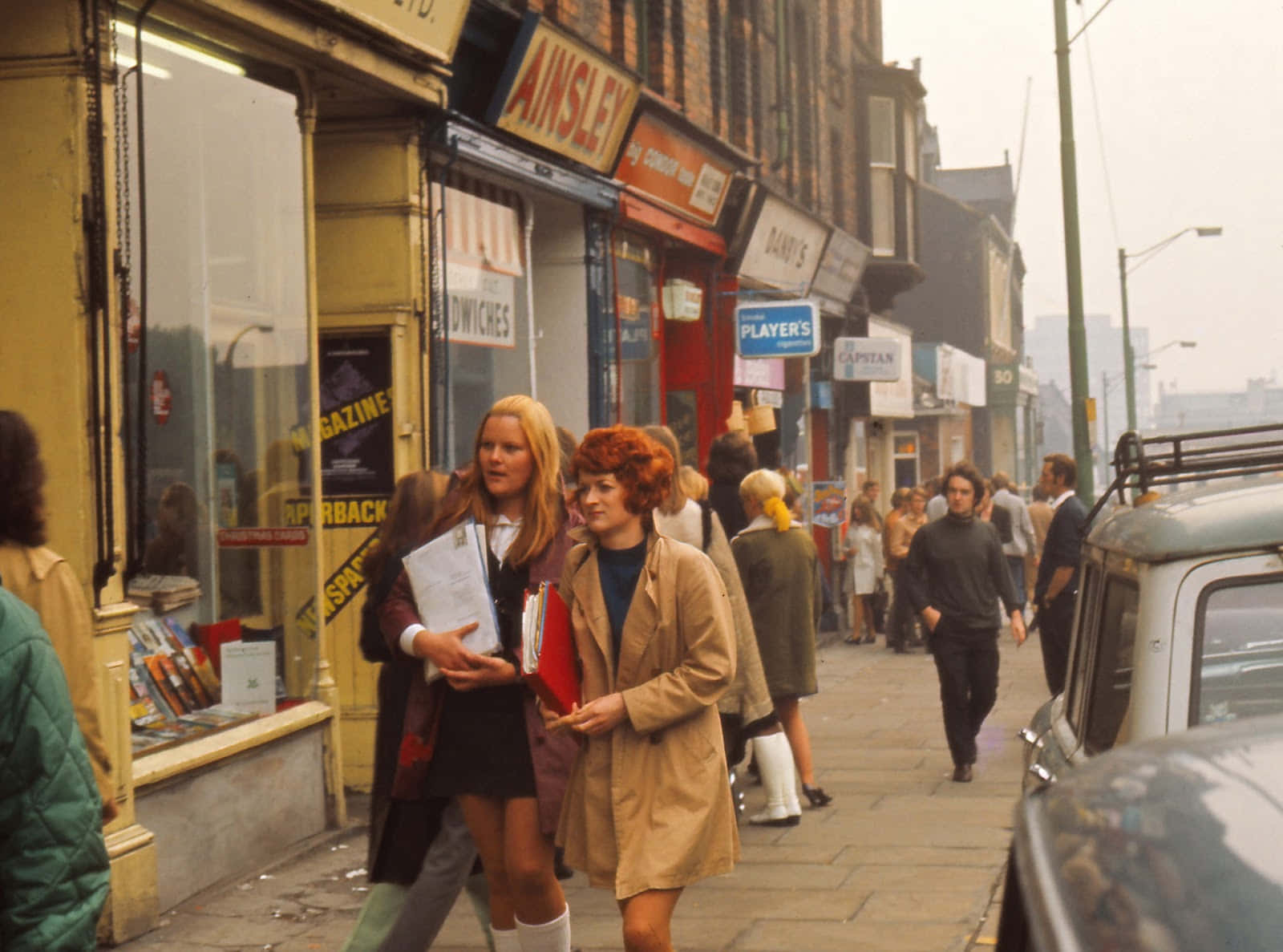 1970年代時尚革命 壁紙 — 兩位女士沿著1970年代英國繁忙的主要街道行走，經過商店和行人，路邊停著復古汽車。