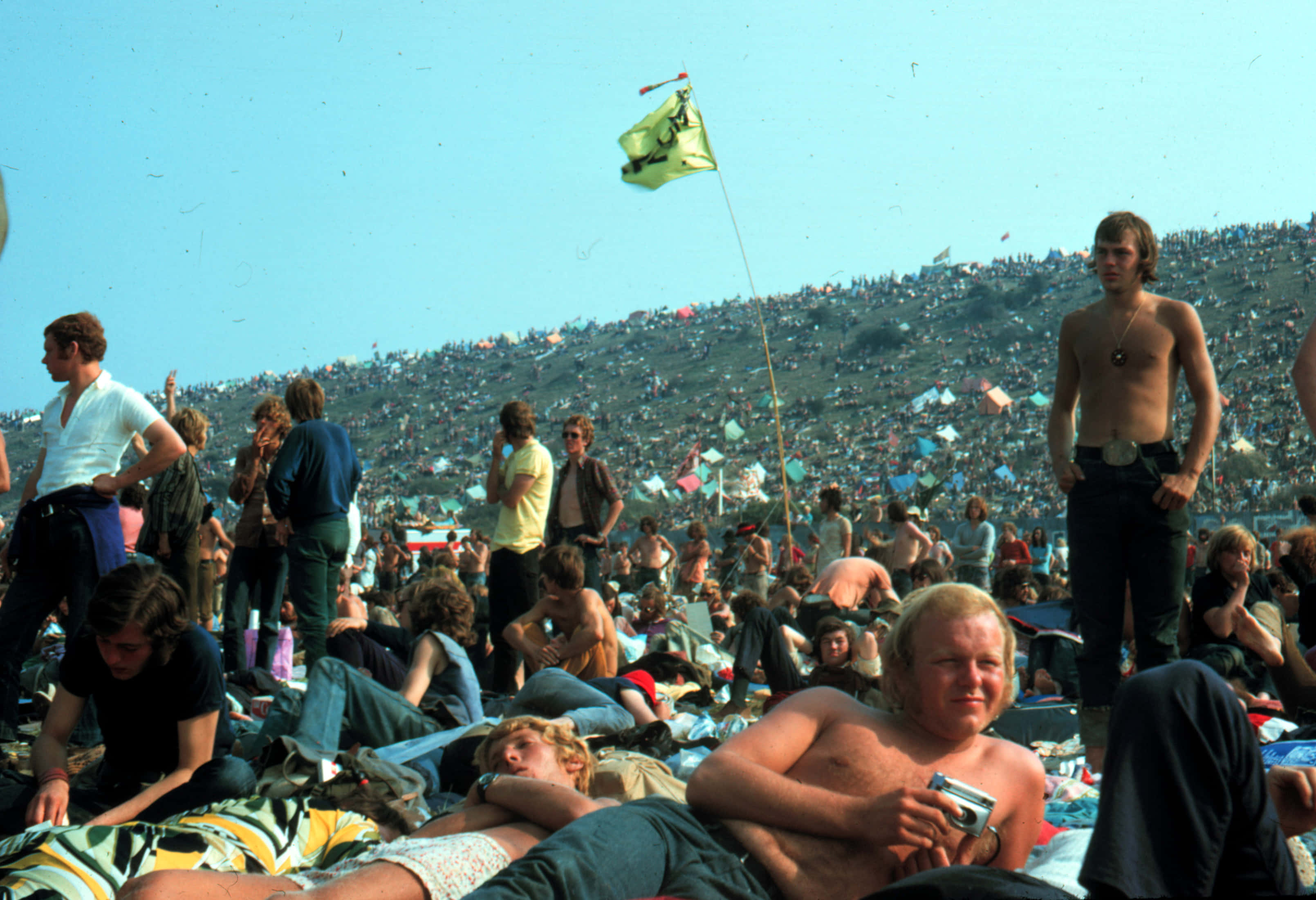 1970年代的生活 壁紙 — 一大群人在晴朗藍天下的大型戶外節日中，在陽光明媚的山坡上放鬆。