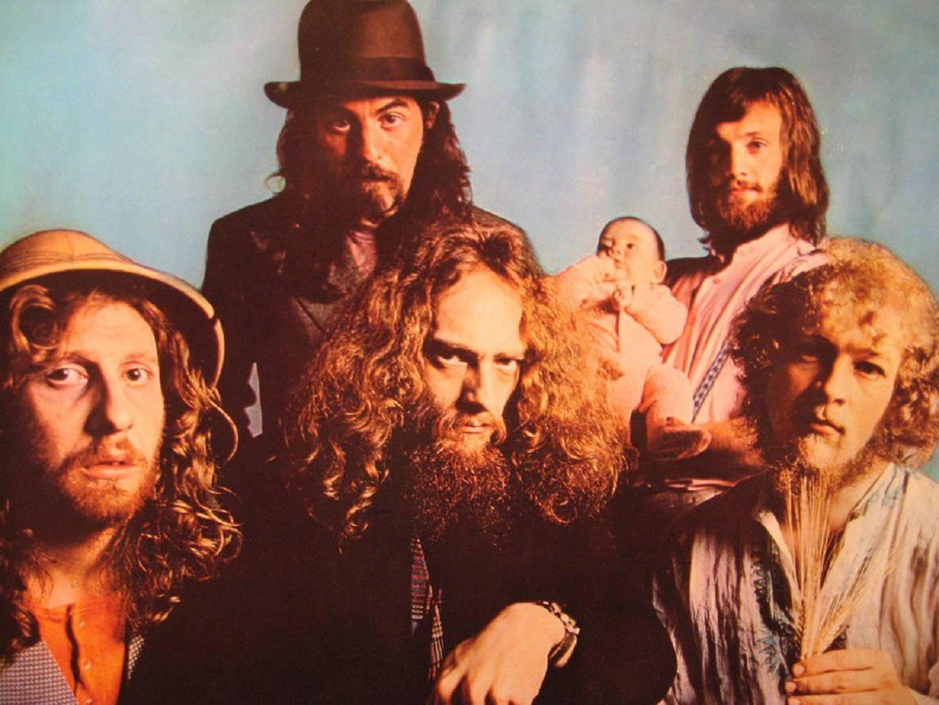 1970年代英國搖滾樂隊Jethro Tull復古影像 壁紙 — 一張復古照片，五位留長髮鬍鬚的男子和一個嬰兒，在溫暖燈光下的藍色背景前擺姿勢。
