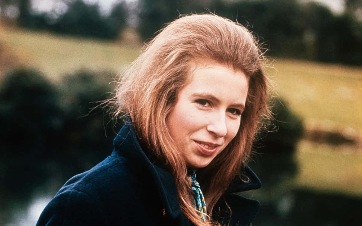 1970年的年輕安妮公主 壁紙 — 一位年輕女性，紅褐色頭髮，穿著深藍色外套，在戶外溫柔微笑。