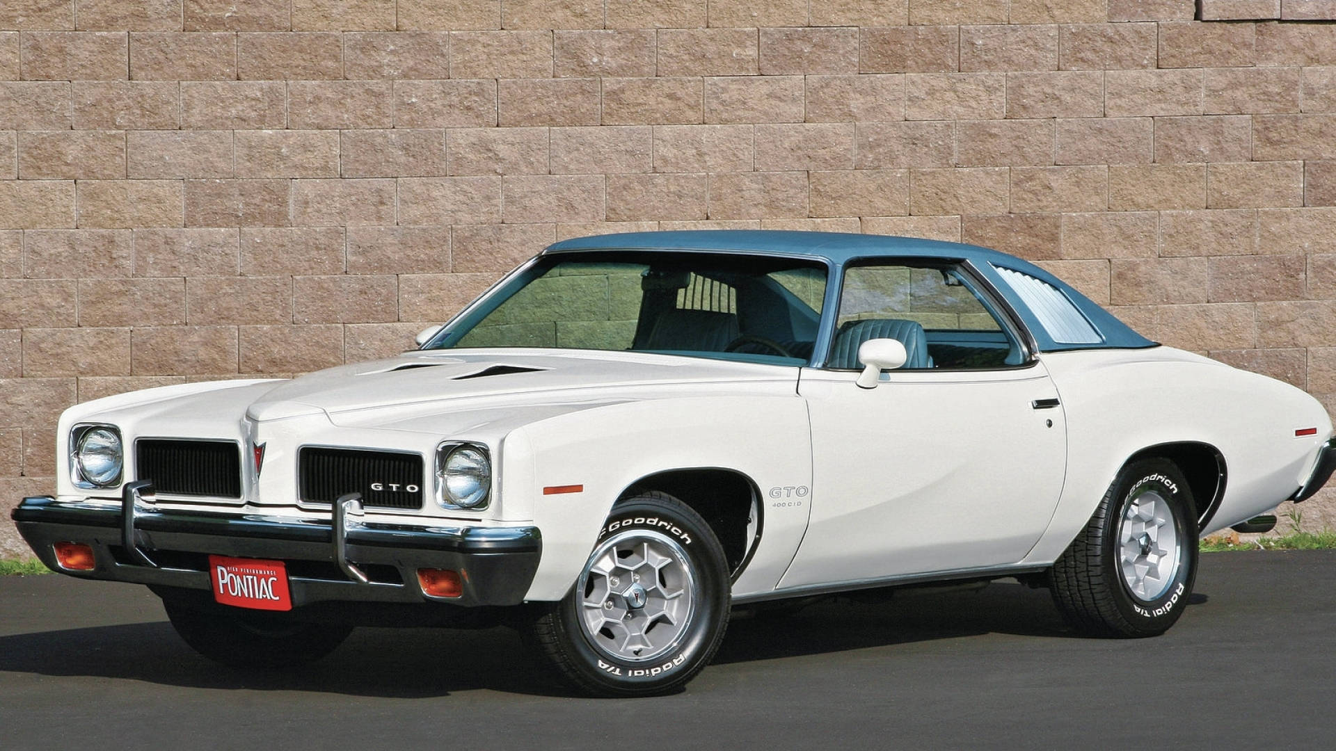 1973 年白色 Pontiac GTO 壁紙 — 一架1970年代白色Pontiac GTO肌肉車，藍色車頂，停泊在石牆旁邊。