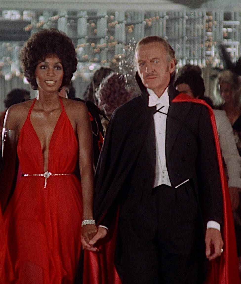 1974年《Vampira》電影大衛·尼文 壁紙 — 一對迷人的情侶穿著正式服裝，手牽手走過一個閃閃發光、賓客滿座的舞廳。