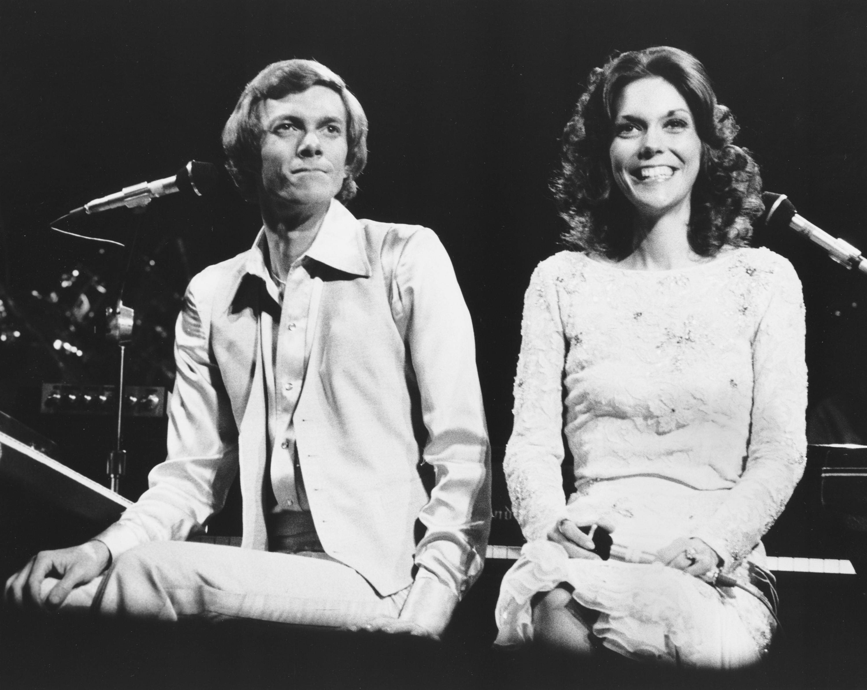 1976年卡朋特兄妹倫敦演出 壁紙 — 一張黑白照片，一對微笑的男女坐在燈光昏暗的舞台上的鋼琴前，旁邊有麥克風。