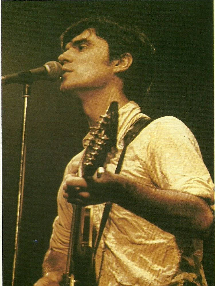 1977 年 Talking Heads 海報攝影 壁紙 — 一位音樂家在溫暖的舞台燈光下，熱情地對著咪高峰唱歌，同時手持電結他。