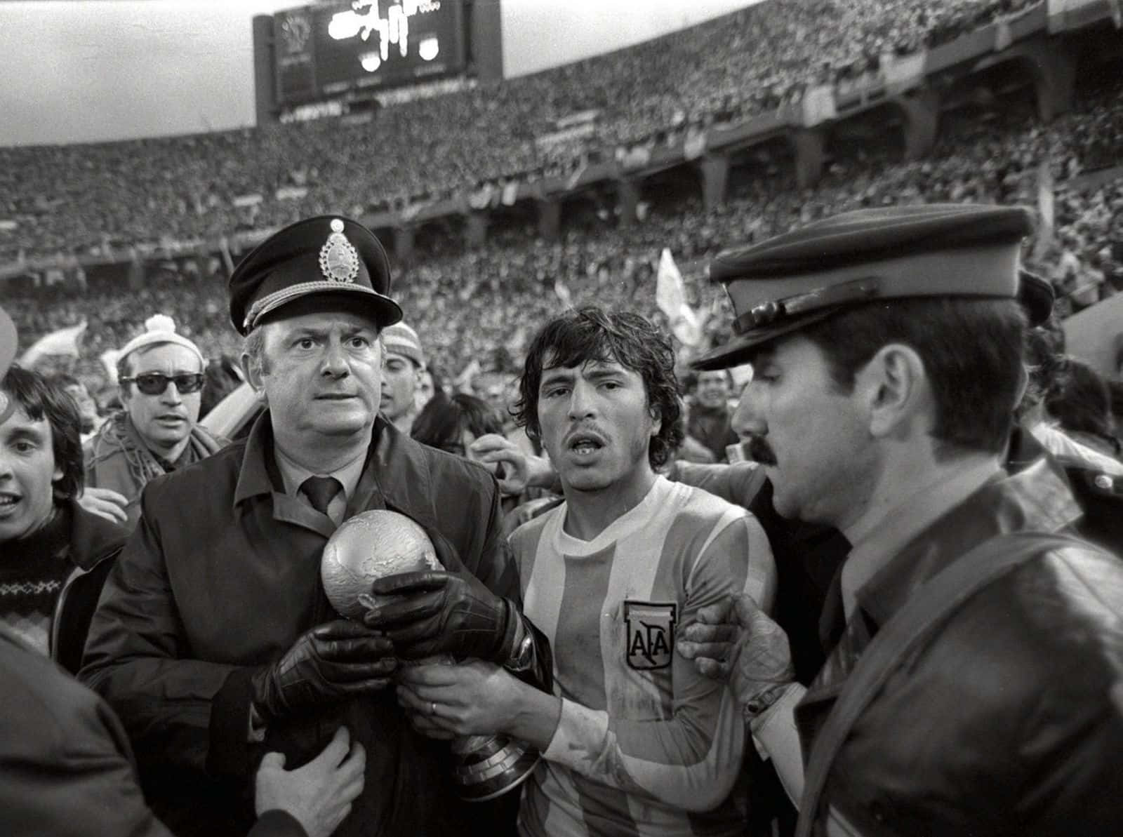 丹尼爾·帕薩雷拉1978年世界盃冠軍 壁紙 — 一張黑白照片捕捉到一位喜悅的阿根廷足球運動員在擠滿觀眾的體育館中，由警察護送下高舉獎盃。