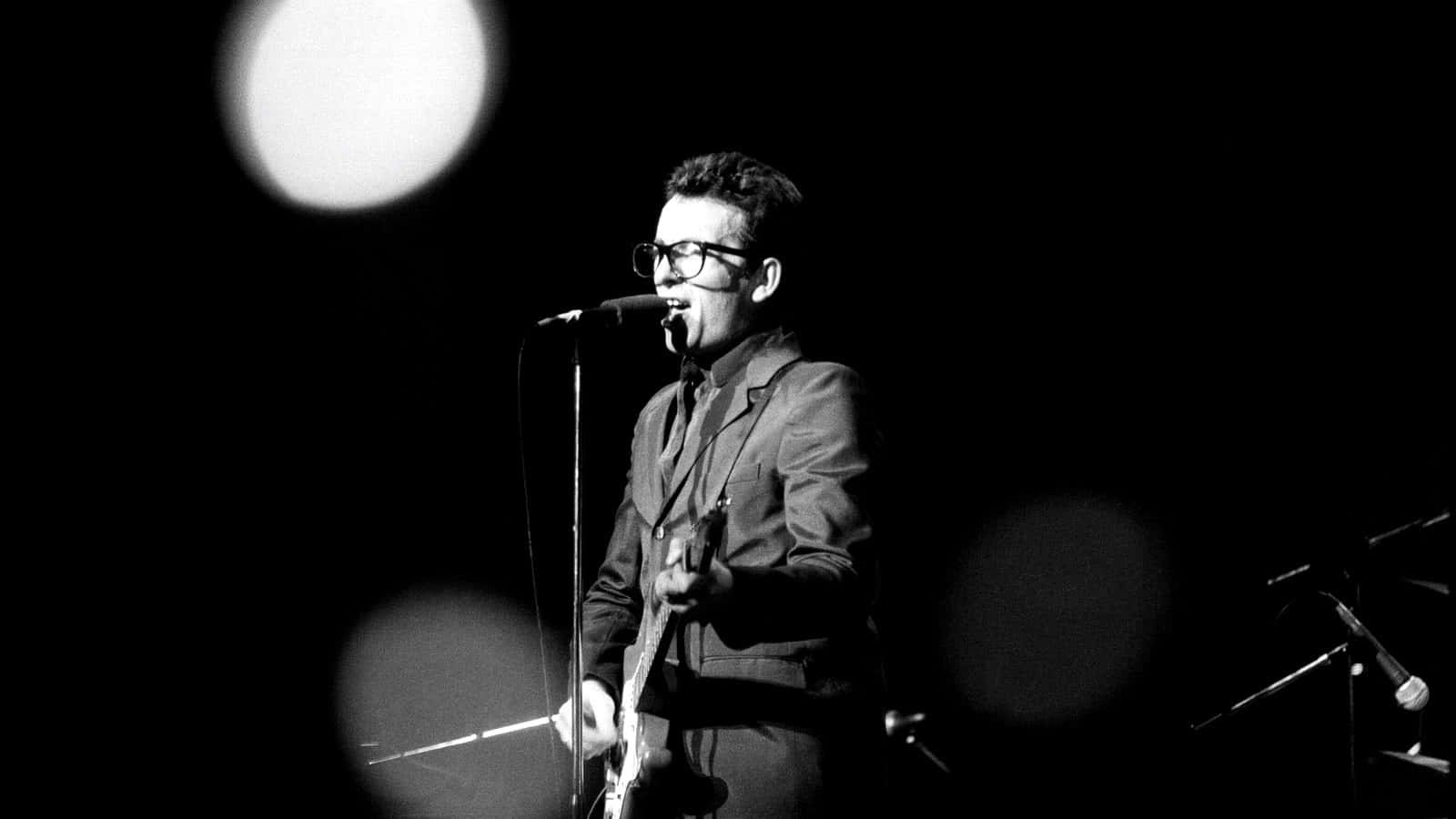 1979年英國歌手Elvis Costello 壁紙 — 一張黑白照片，一名戴眼鏡的男子在舞台燈光下拿著結他對著咪高峰唱歌。