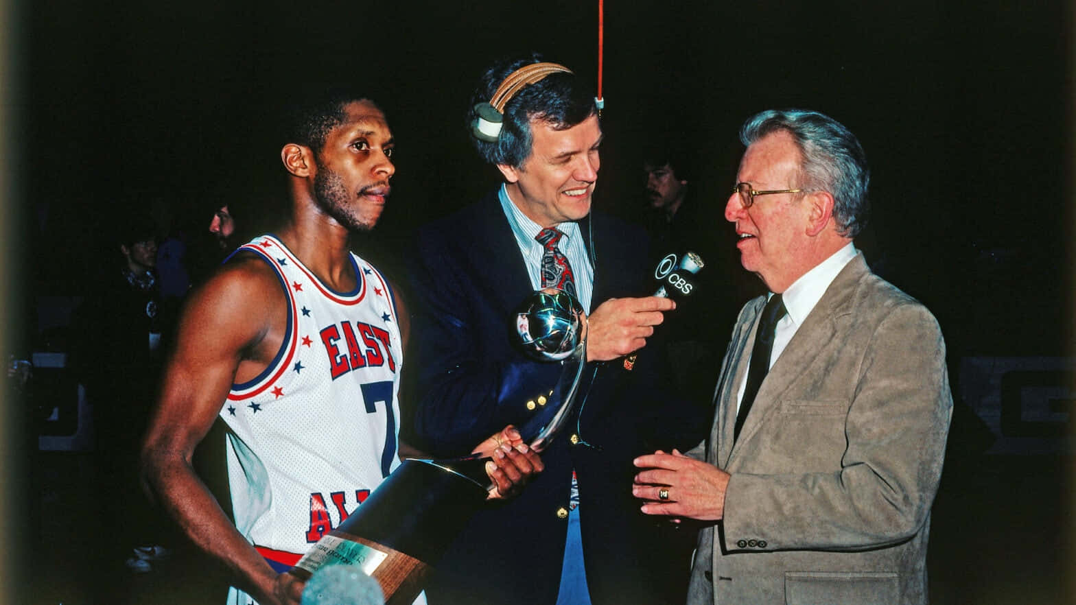 1981年NBA全明星賽MVP 奈特·阿奇巴爾德 壁紙 — 一名身穿 'EAST' 球衣的籃球運動員手持獎盃，在昏暗的活動場景中被兩名男子用咪高峰採訪。
