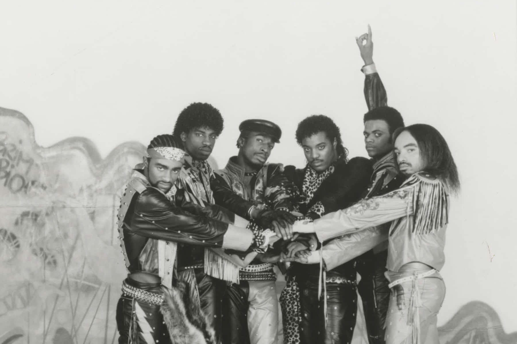1984 Grandmaster Flash 和 The Furious Five 壁紙 — 六個穿著大膽1980年代時尚的男人，在一個引人注目的黑白照片中，緊密相連地對著塗鴉牆擺姿勢。