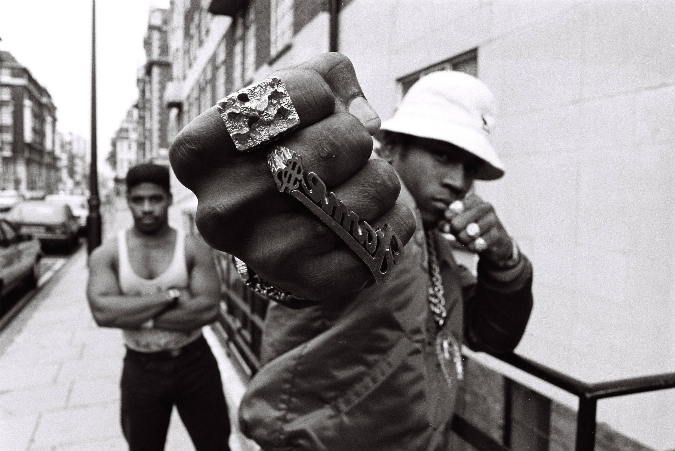1986 年 LL Cool J 展示金戒指 壁紙 — 一張黑白照片，展示一個男人緊握的拳頭，上面裝飾著大戒指和鏈條，站在城市街道上，背景有另一個男人。