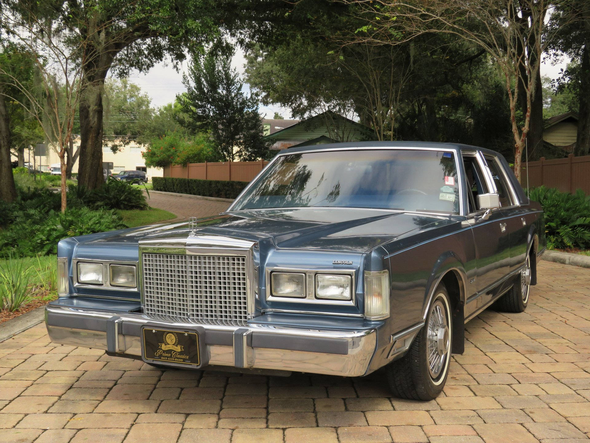 1986 林肯 Town Car 停泊於鋪砌門前 壁紙 — 一輛經典藍色林肯大陸轎車在日光下閃耀，停放在磚砌車道上，周圍環繞著茂密的綠樹和郊區房屋。
