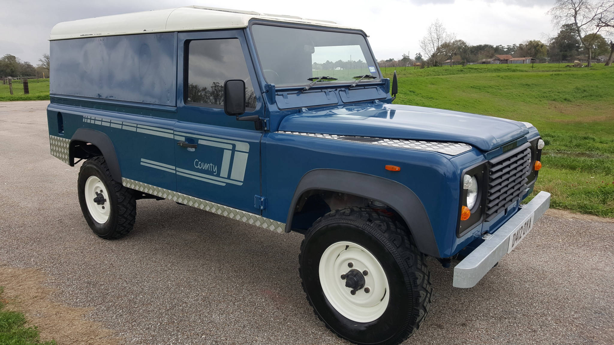 1987款路虎衛士 iPhone 主題壁紙 壁紙 — 一輛經典藍色 Land Rover Defender County 客貨車停泊在碎石路上，旁邊是草地，天空多雲。