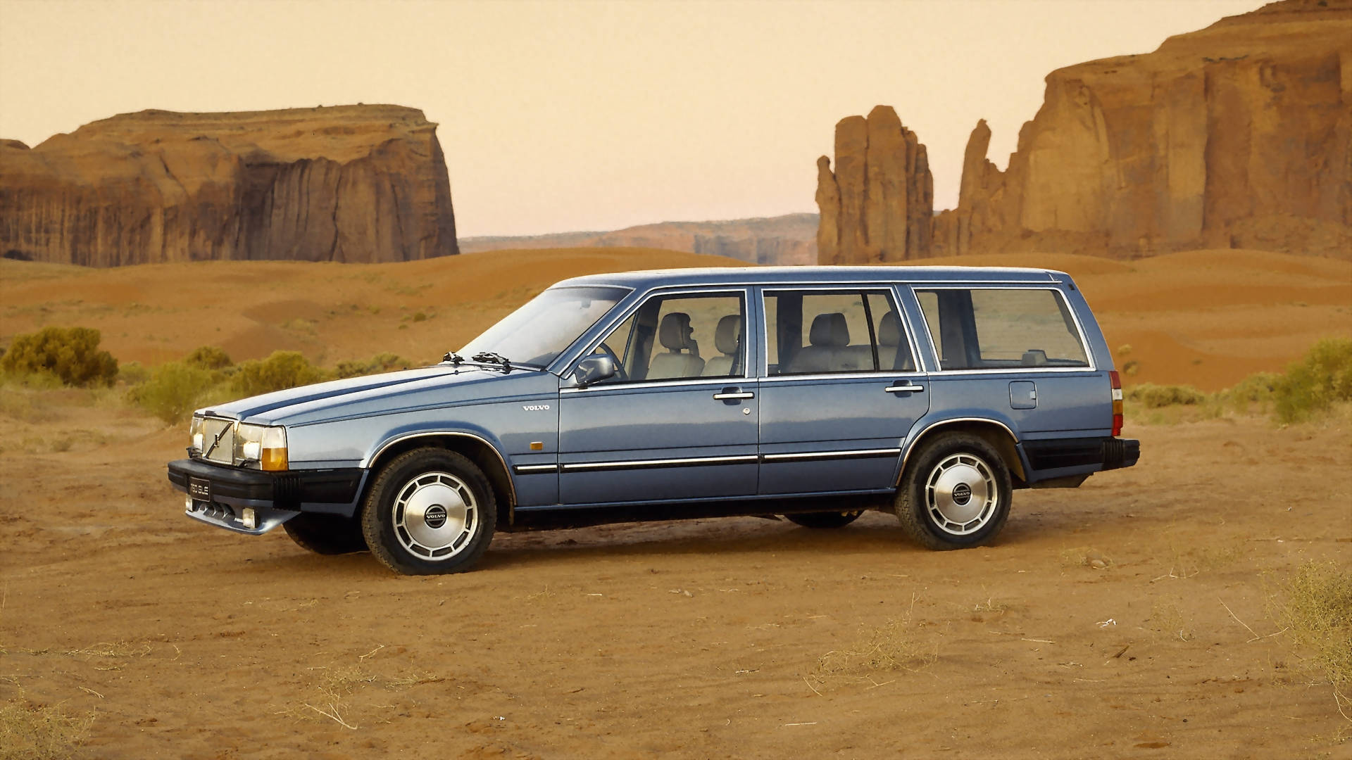1992款740 Turbo沃爾沃iPhone照片 壁紙 — 一輛藍色富豪旅行車停在佈滿灰塵的地形上，背景是朦朧天空下高聳的紅色岩層。