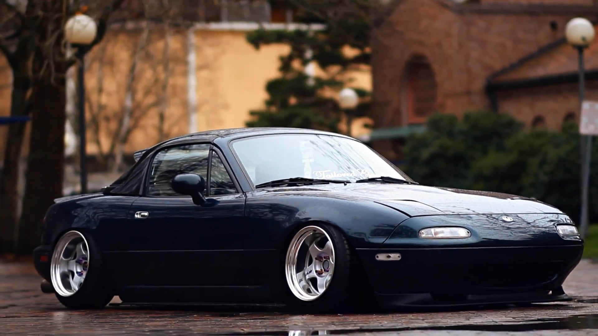 1995年深藍色 Mazda MX-5 Miata 壁紙 — 一架時尚深藍色改裝Miata跑車停泊在濕滑的鵝卵石街道上，背景是模糊的城市景觀。
