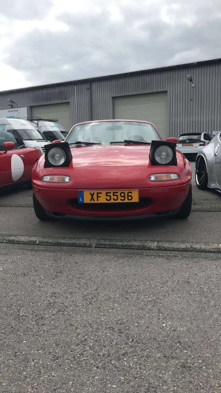 1995年馬自達MX-5 Miata紅色跑車 壁紙 — 一架鮮紅色經典開篷跑車停泊在瀝青路上，背景是工業建築物，天空多雲。