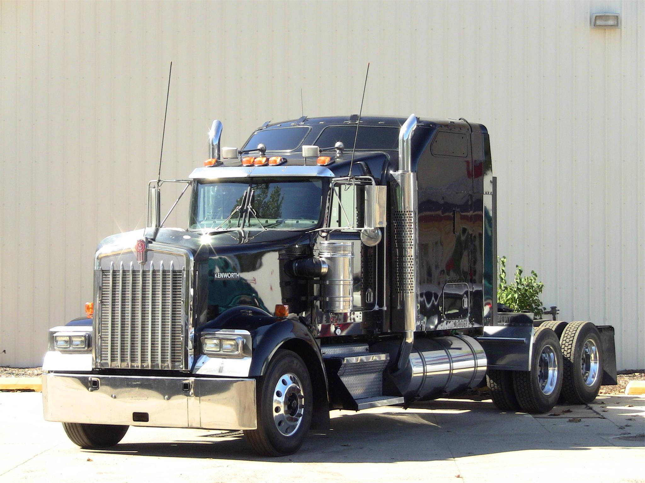 1996年肯沃斯W900 壁紙 — 一輛閃亮的黑色Kenworth半掛卡車，帶有鍍鉻裝飾，停在米色金屬建築前，陽光明媚。