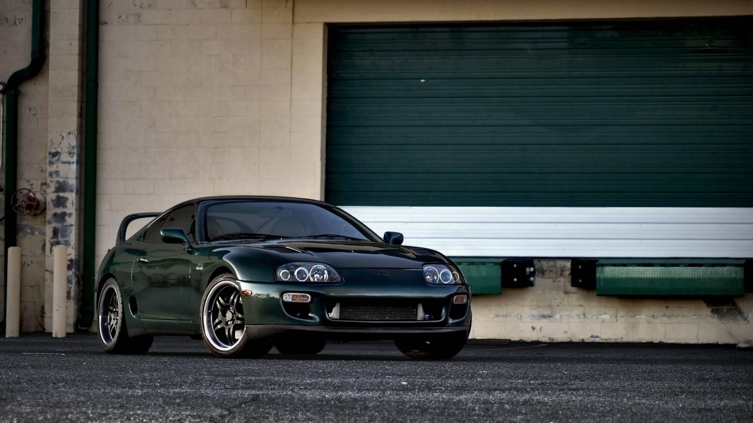 1997 年 Toyota Supra Mk4 深寶石綠珍珠色 壁紙 — 一架時尚的深綠色豐田Supra跑車停泊在帶有綠色捲簾門的倉庫前。