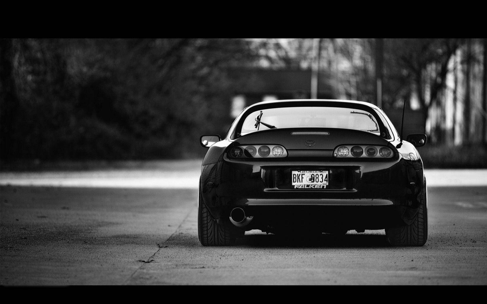 黑白1998年豐田Supra Mk4 壁紙 — 從後方觀看，一輛時尚黑色跑車停在寧靜的道路上，背景樹木以單色模糊處理。