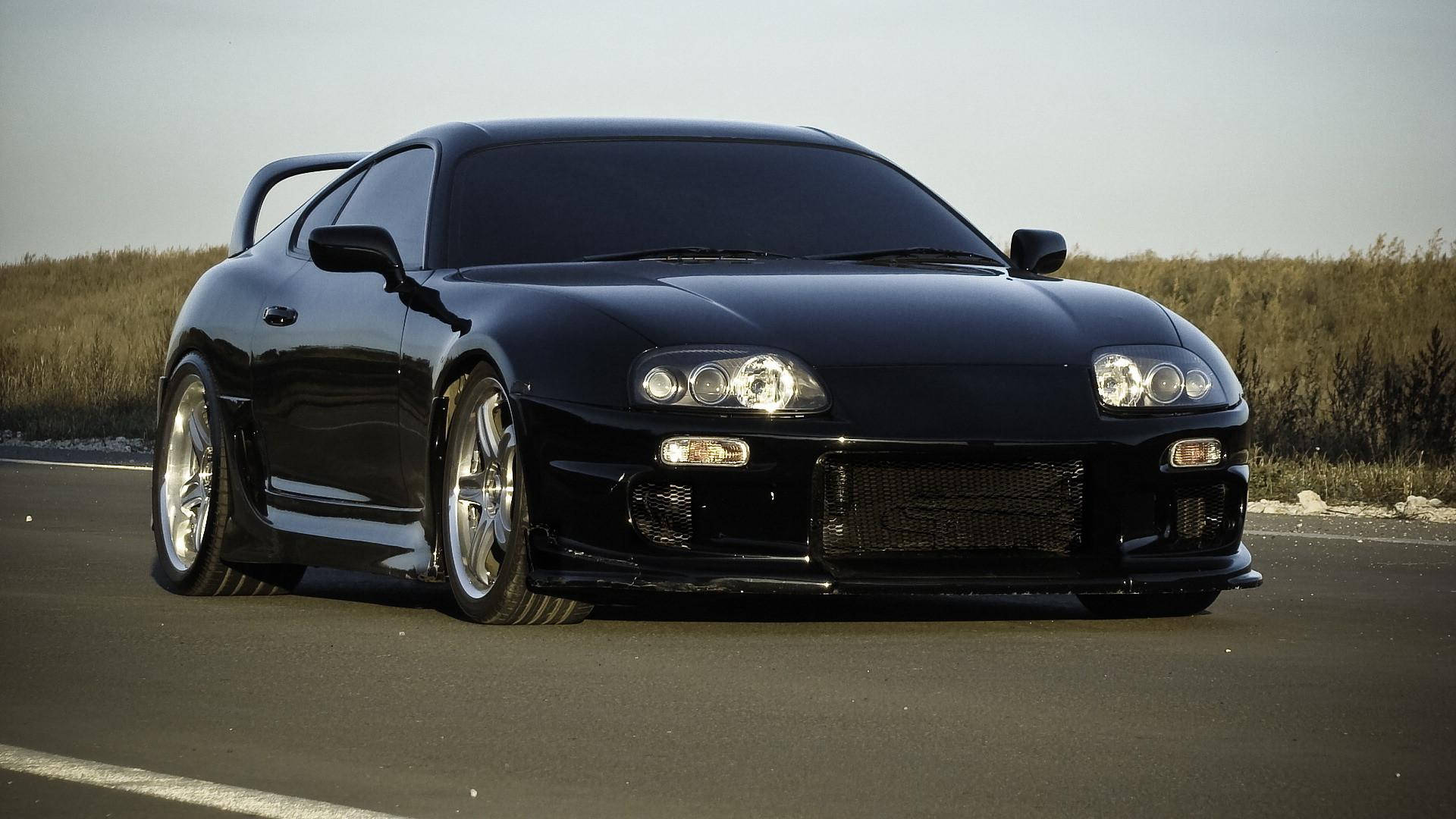 1998年黑色Toyota Supra Mk4 壁紙 — 一架時尚黑色跑車配備定制輪圈和後擾流板，停泊在空蕩蕩的道路旁，旁邊是乾草，天空朦朧。