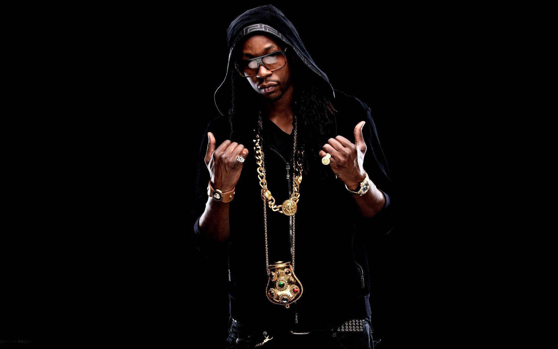 2 Chainz 海報造型 壁紙 — 一名身穿黑色連帽衫、戴眼鏡的男子，在飾有金鏈和戒指的黑色背景前自信地擺姿勢。