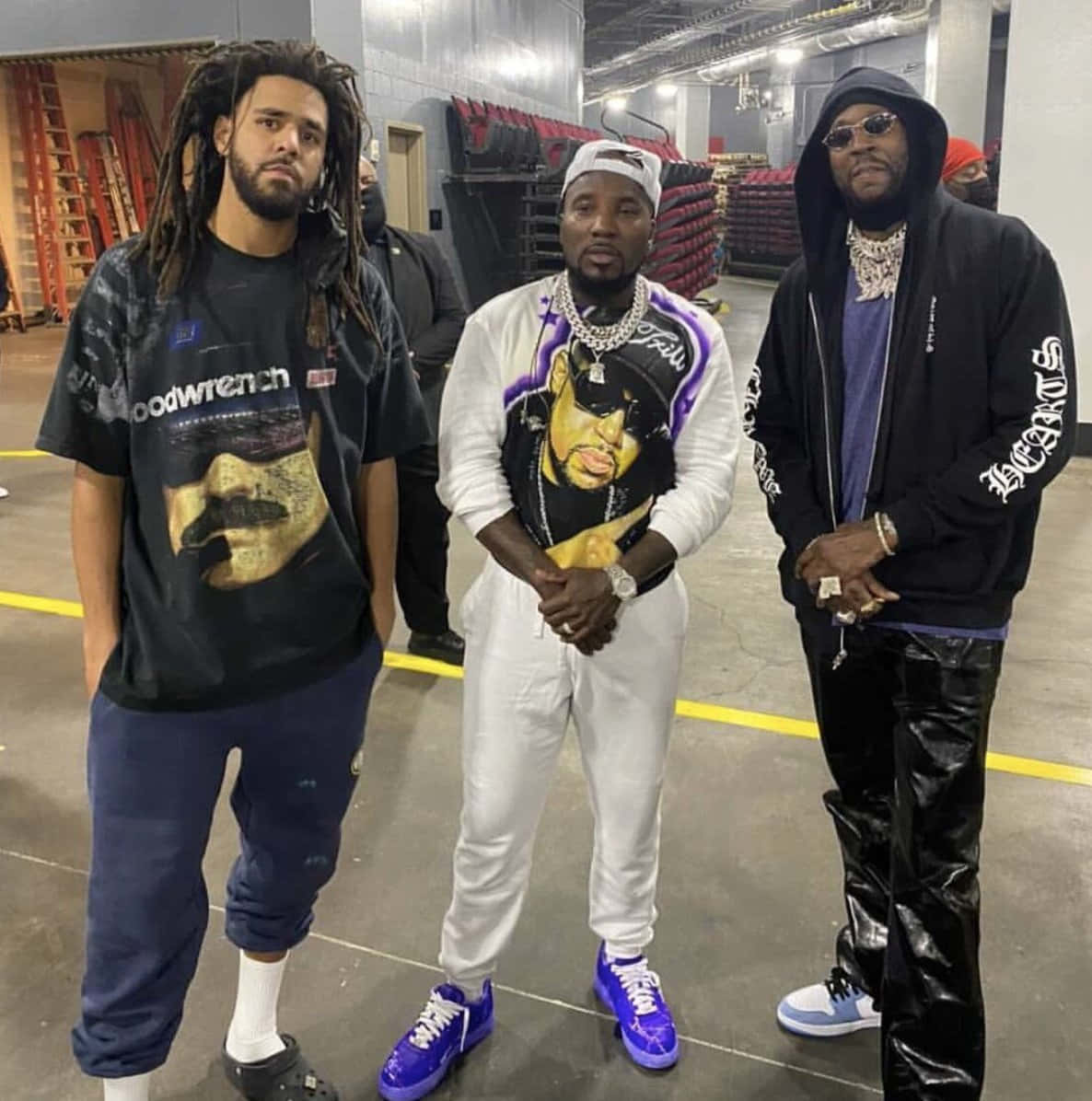 2 Chainz 享受當下時光 壁紙 — 三位時尚男士穿著大膽街頭服飾，在後台倉庫中自信擺姿勢，背景有堆疊的紅色椅子和工業照明。