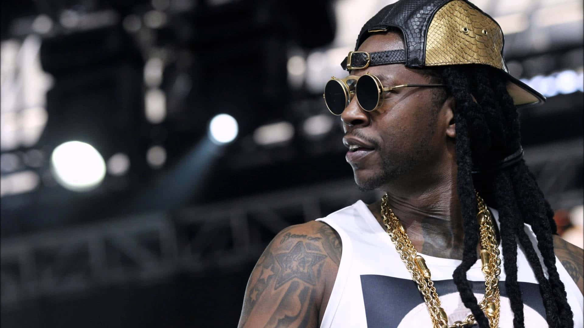 2 Chainz 展現他迷人的笑容 壁紙 — 一位時尚的饒舌歌手，留著辮子頭，戴著金鏈，自信地站在燈光昏暗的演唱會舞台上。