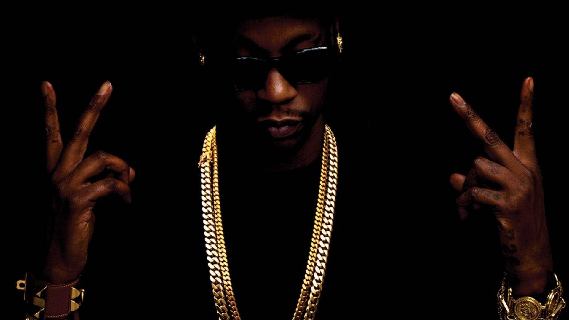 2 Chainz 戲劇性攝影 壁紙 — 一名戴著太陽眼鏡和金鏈的男子在黑色背景上以戲劇性燈光做出和平手勢。