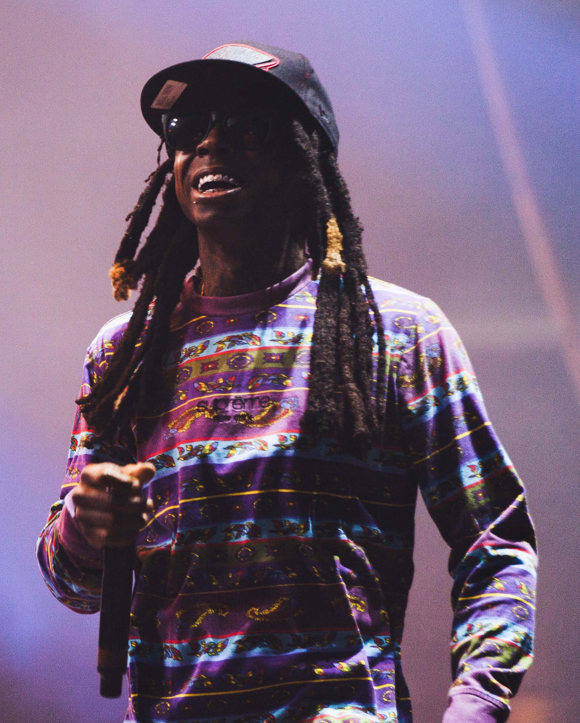 2 Chainz 留下片刻 壁紙 — 一位微笑的表演者，留著辮子頭、戴著太陽眼鏡，在紫色舞台燈光下對著麥克風唱歌，穿著色彩繽紛的圖案襯衫。