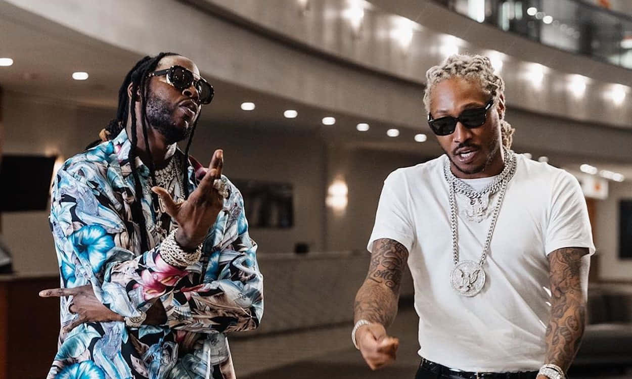 2 Chainz 讓音樂說話 壁紙 — 兩位時尚的饒舌歌手戴著太陽眼鏡，在溫暖燈光的現代室內大堂自信地做手勢。