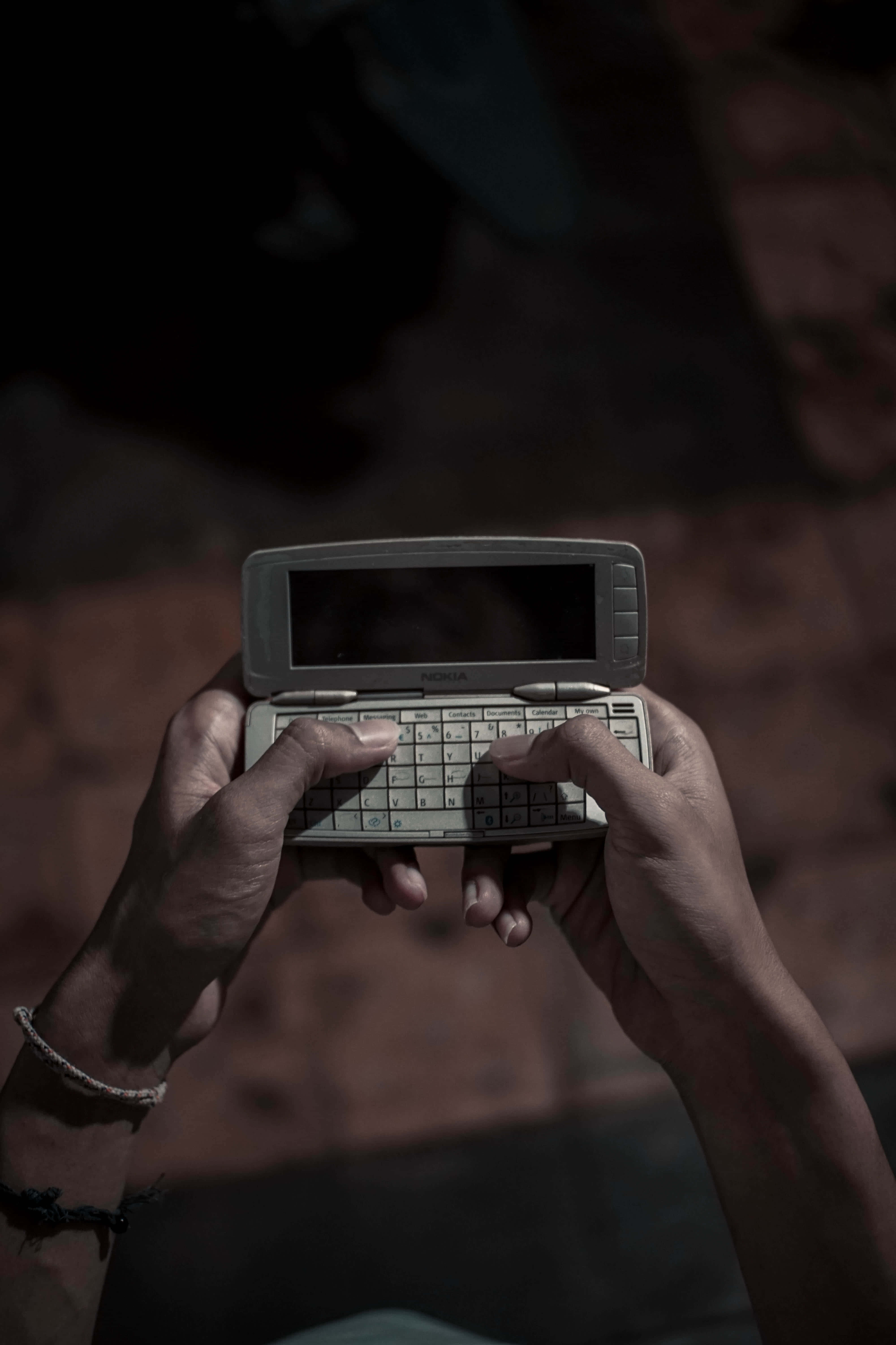 2000 年代 Nokia 93000 壁紙 — 雙手在昏暗燈光下，手持一部帶鍵盤的復古銀色翻蓋手機，背景為模糊的微紅色。
