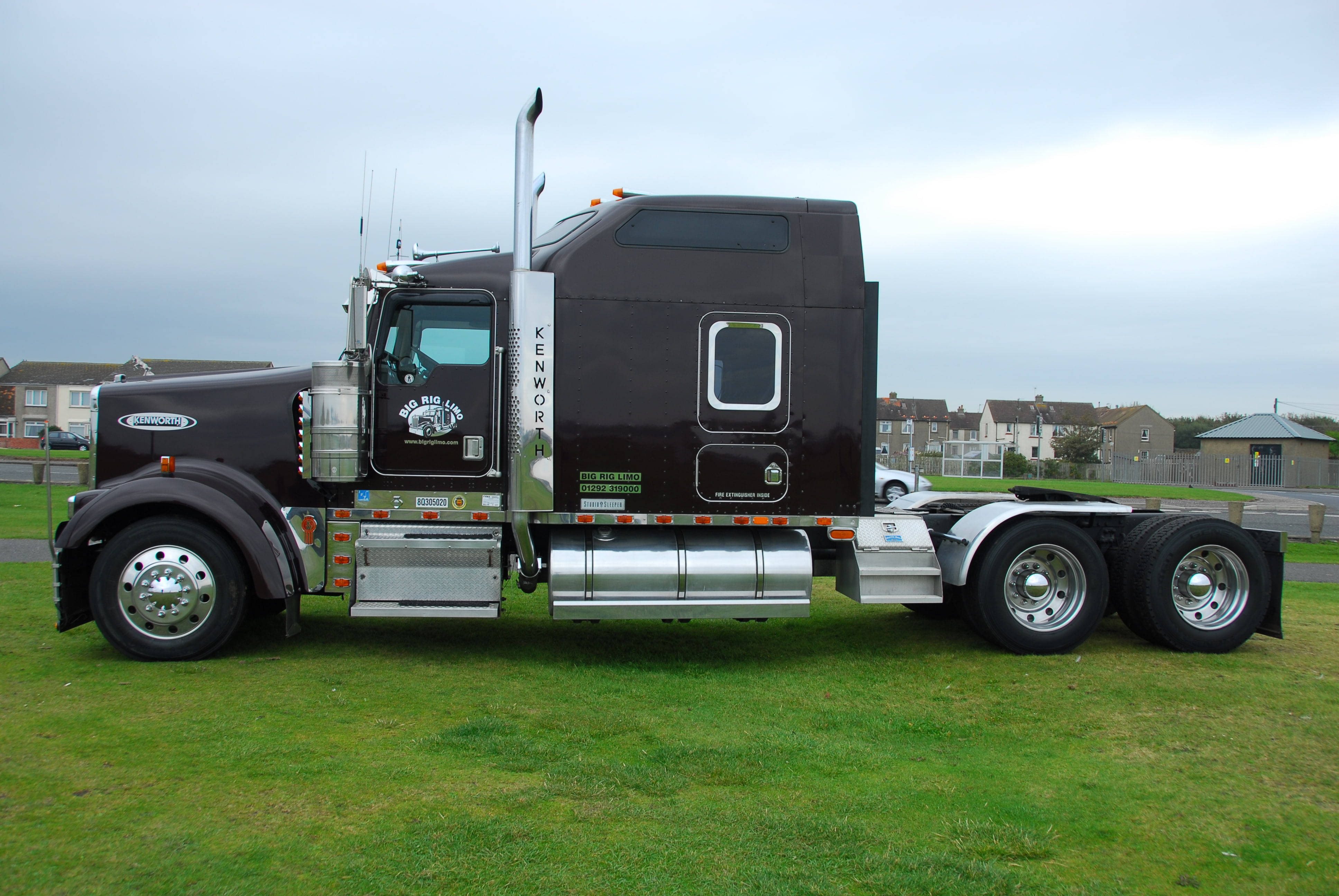 2000 年肯沃斯 W900 卡車 壁紙 — 一架時尚的黑色Kenworth半掛卡車帶有鍍鉻裝飾，停泊在陰天下的草地上。