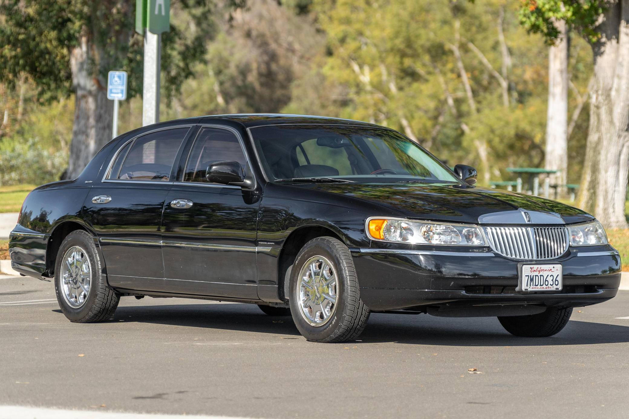 2001 林肯 Town Car 停泊於停車場 壁紙 — 一架時尚的黑色林肯Town Car停泊在陽光明媚的街道上，背景有樹木和野餐桌。