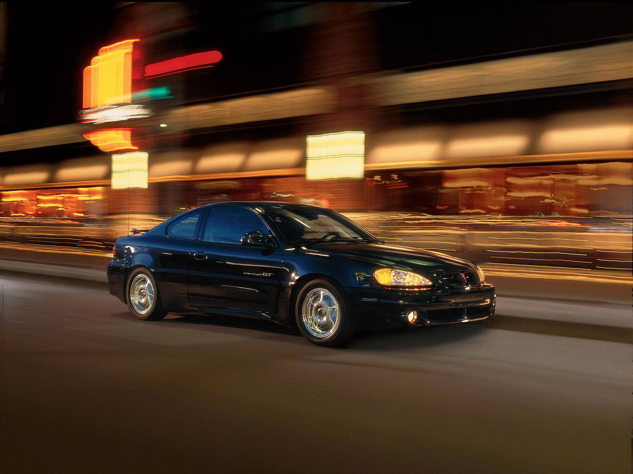 2002龐蒂克Grand GT 壁紙 — 一架型格黑色Pontiac Grand Am GT在夜晚霓虹燈閃爍的街道上飛馳，背景帶有動態模糊效果。